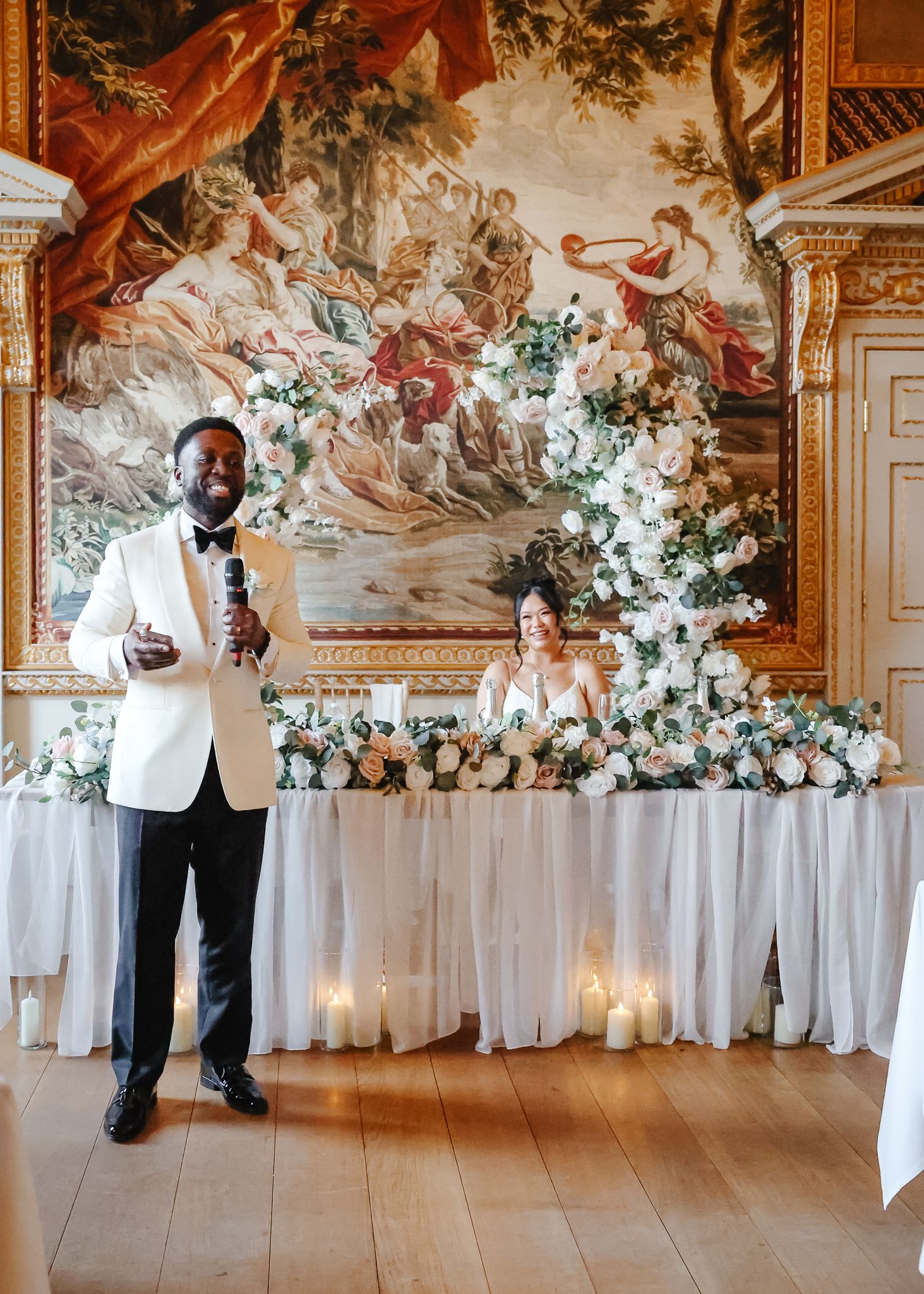 Elegant wedding reception setup with white linens and floral arrangements in grand ballroom with historic murals.