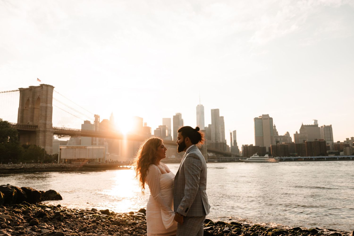 Couple stands together near Brooklyn Bridge at sunset with Manhattan skyline reflecting on the water.