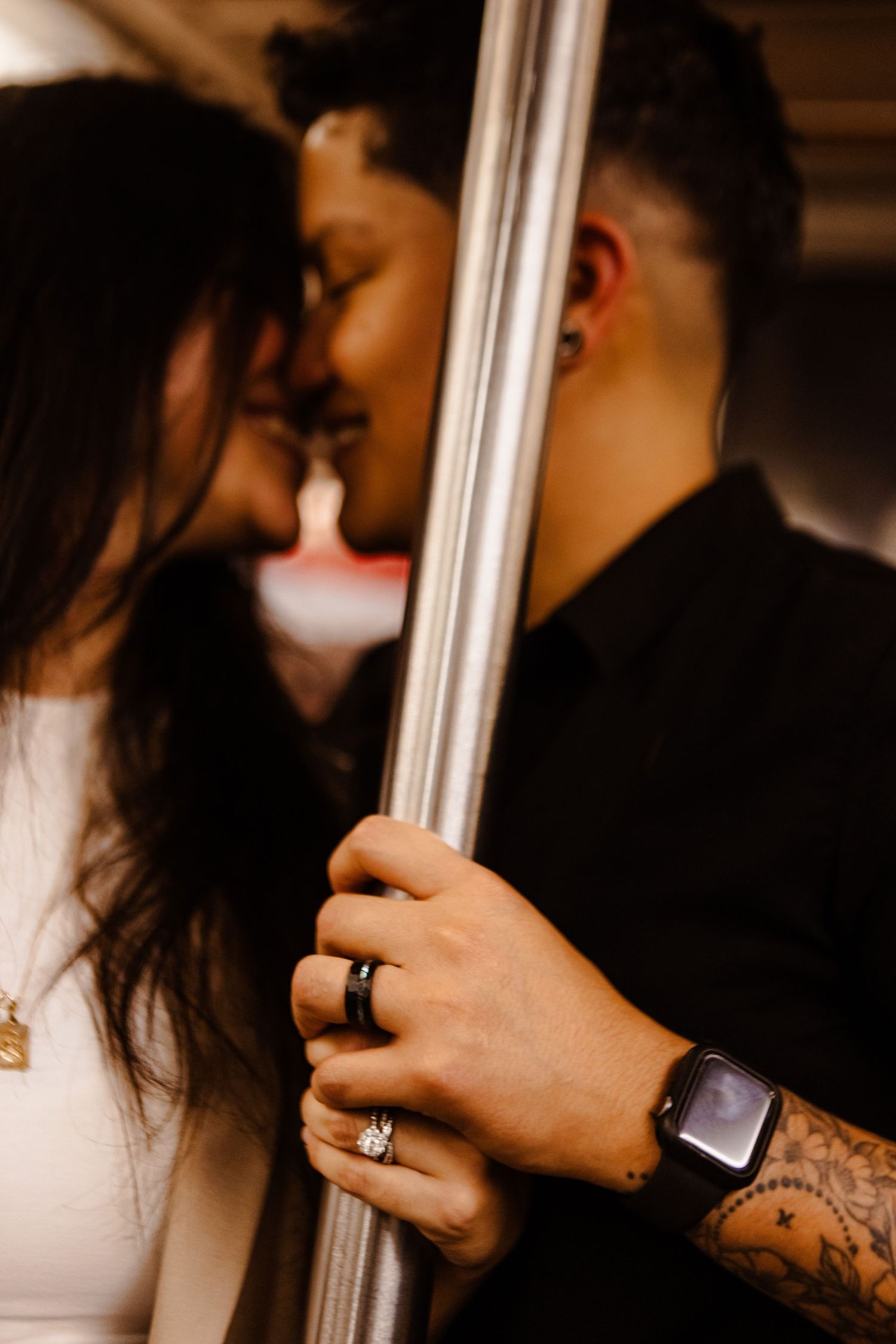 Close-up of couple nearly kissing on subway, showing engagement ring and tattooed hands on pole.