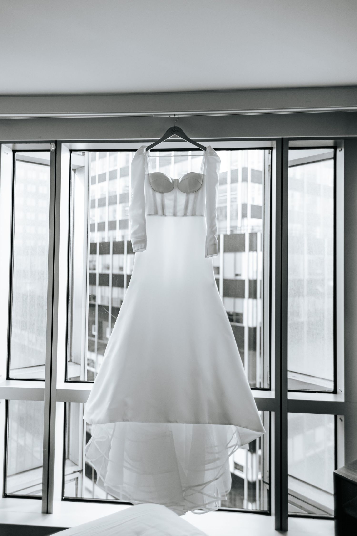Elegant white wedding dress with lace sleeves hanging by a window with city views.