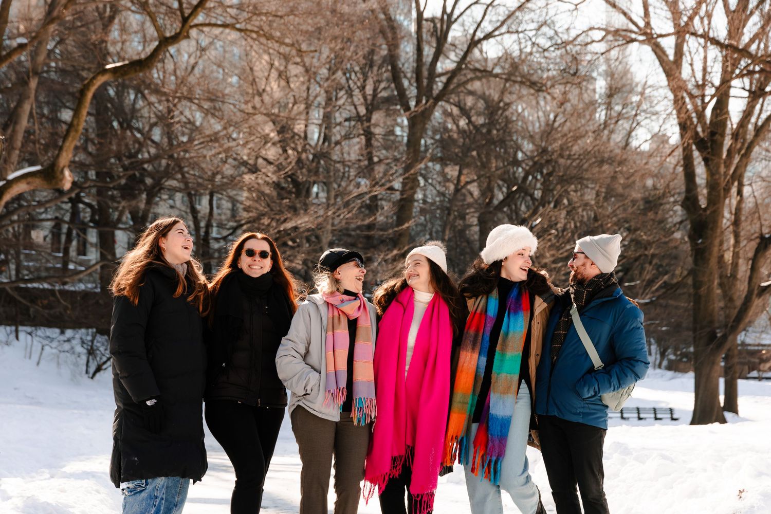 Six women laughing and walking together through a snowy Central Park wearing vibrant winter scarves and coats.