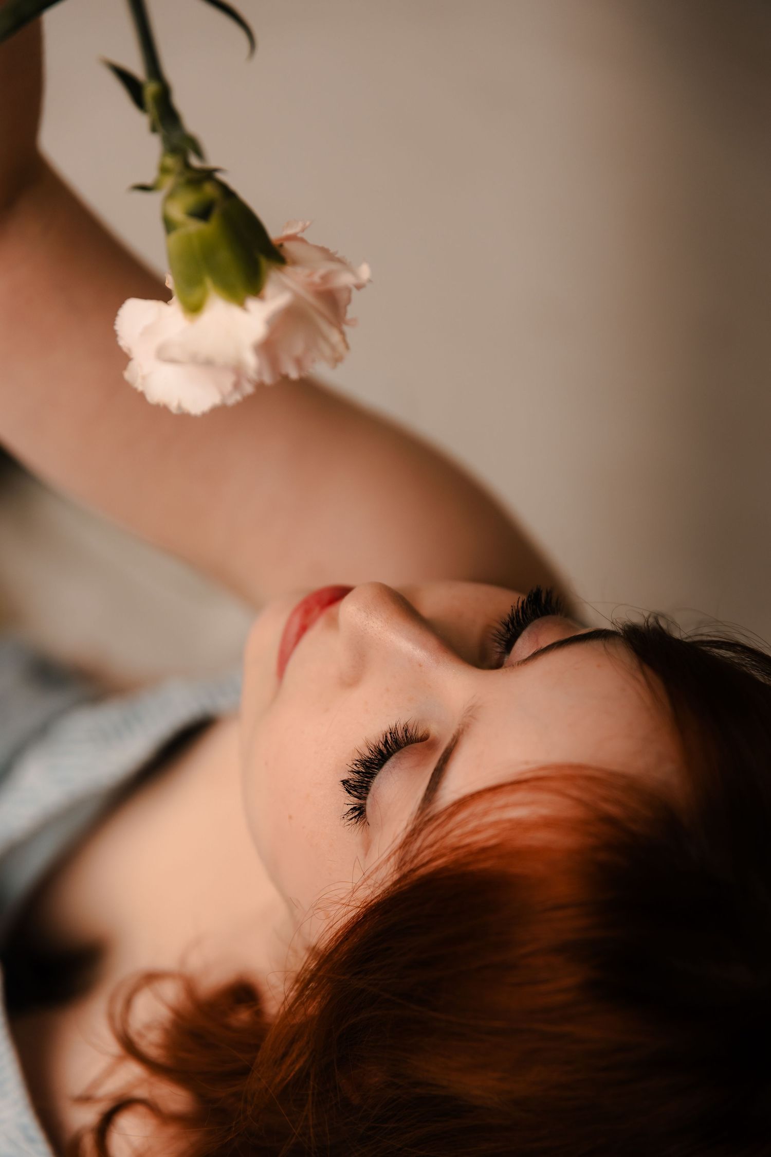 Close-up of a redhead woman lying down with eyes closed, holding a white carnation flower above her face.
