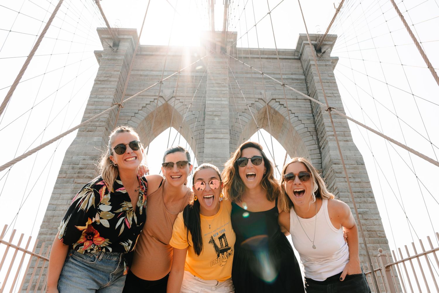 Five smiling women posing together at Brooklyn Bridge tower with sunlight glowing behind them.