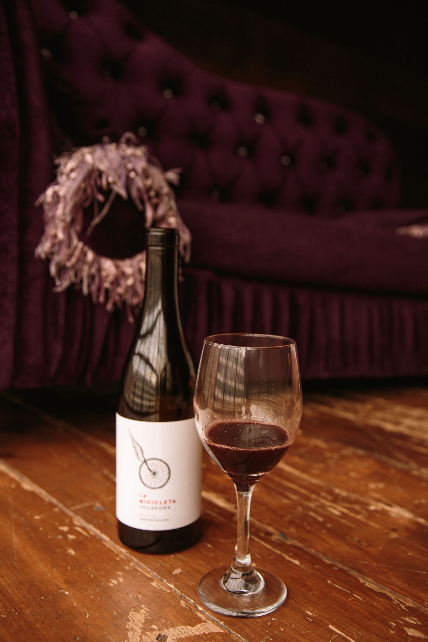 Bottle of La Bicicleta red wine with glass on rustic hardwood floor near purple velvet sofa.