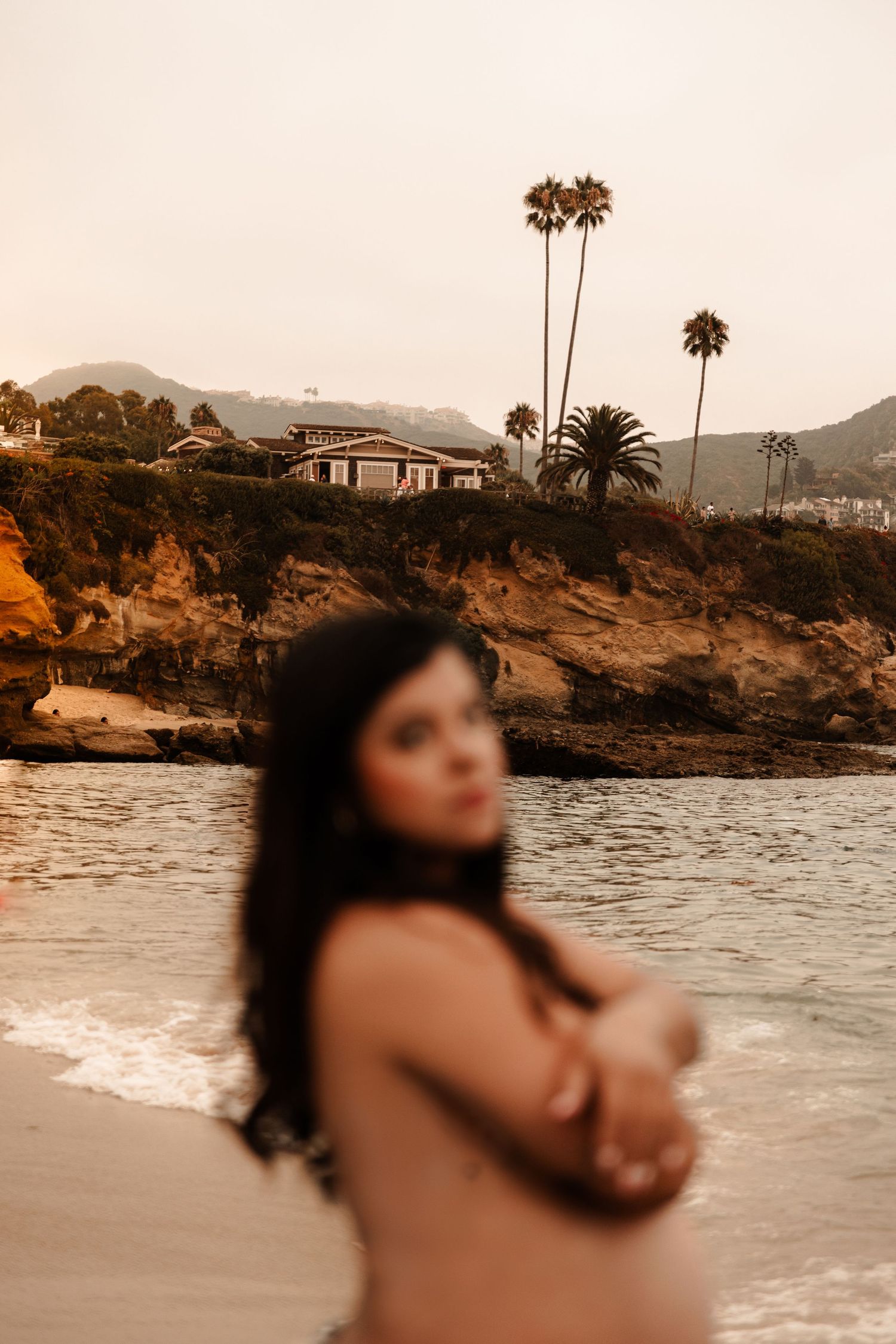 Pregnant woman poses on a California beach at golden hour with palm trees and cliffs in the background.
