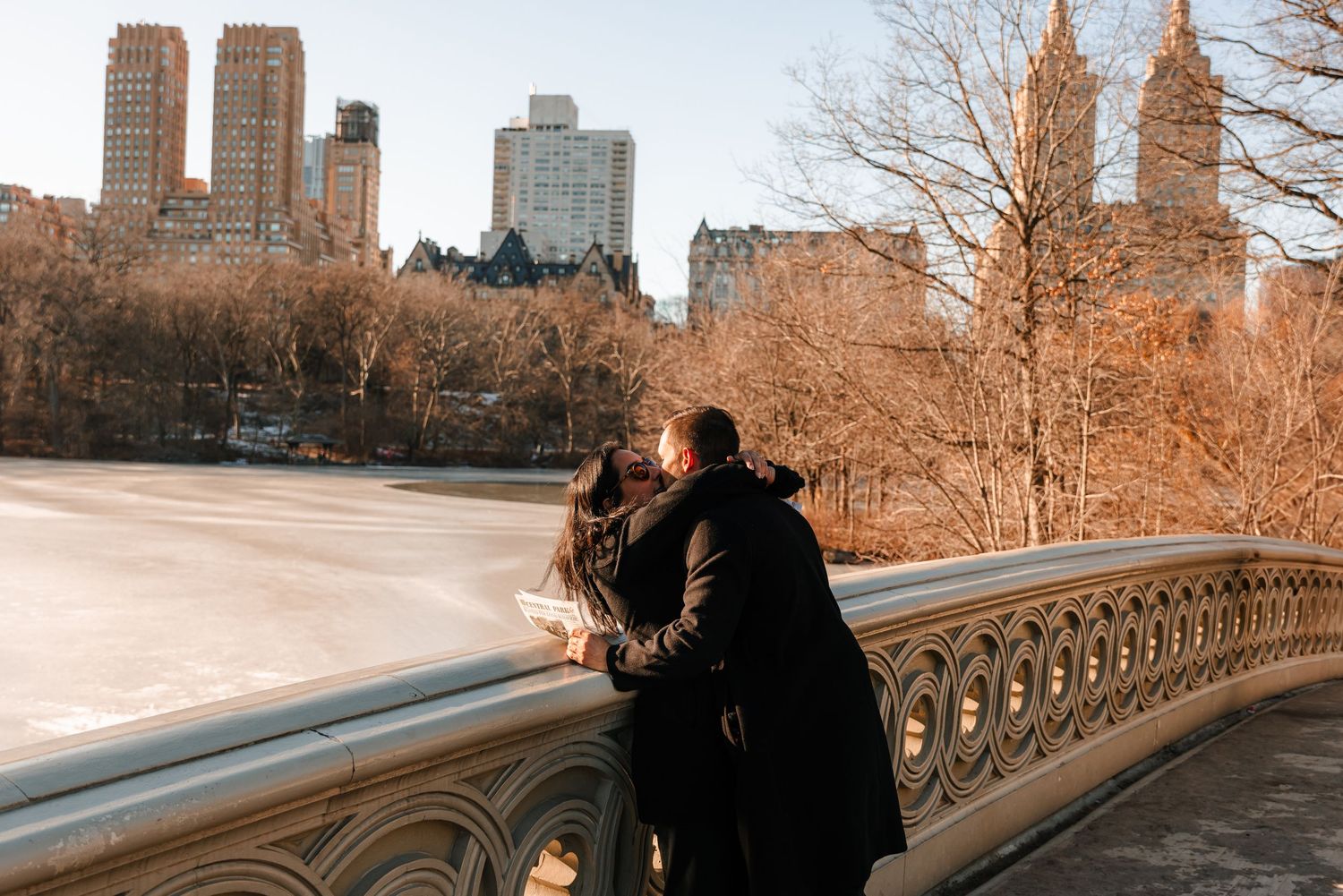 Couple leans on Bow Bridge railing in Central Park overlooking a frozen lake in winter.