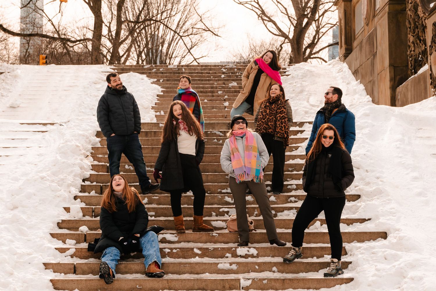 Group of eight friends posing playfully on snowy stone steps in a winter park setting.