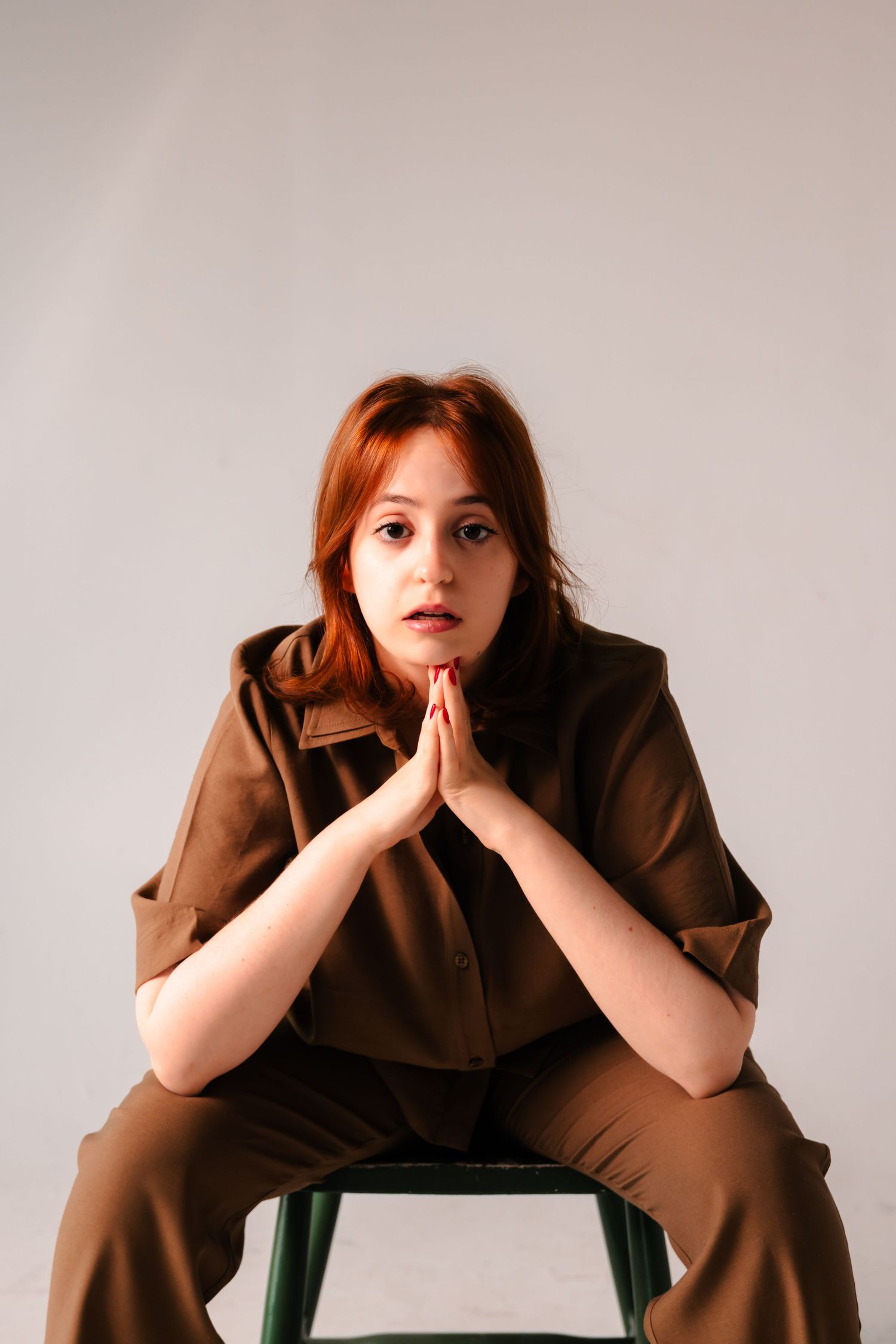 Young redhead woman in brown outfit sits on stool with hands pressed together in prayer pose, looking directly at camera.