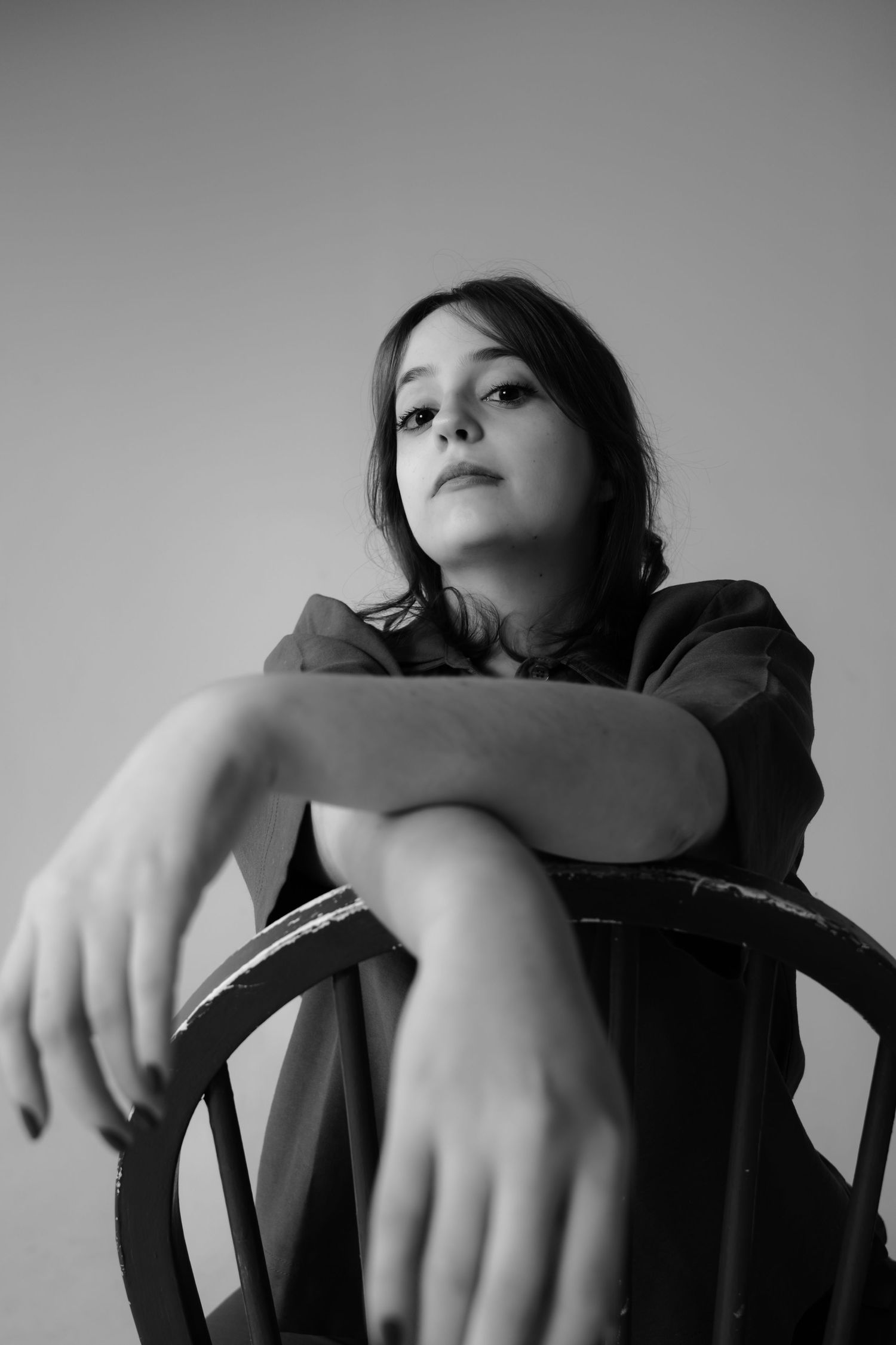 Black and white portrait of a young woman sitting backwards on a wooden chair, gazing upward with a contemplative expression.