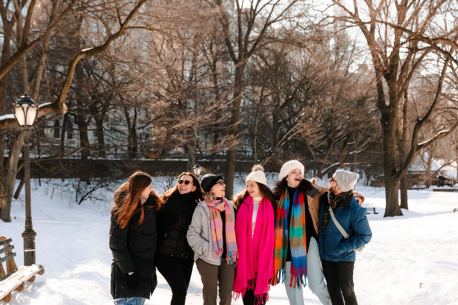 Six smiling women walking together arm in arm through a snow-covered Central Park in colorful winter attire.