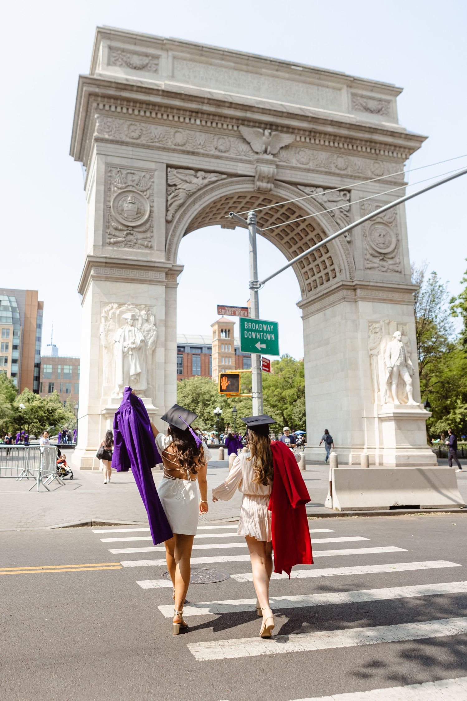 Two graduates in purple and red robes walk toward Washington Square Arch in NYC crosswalk.