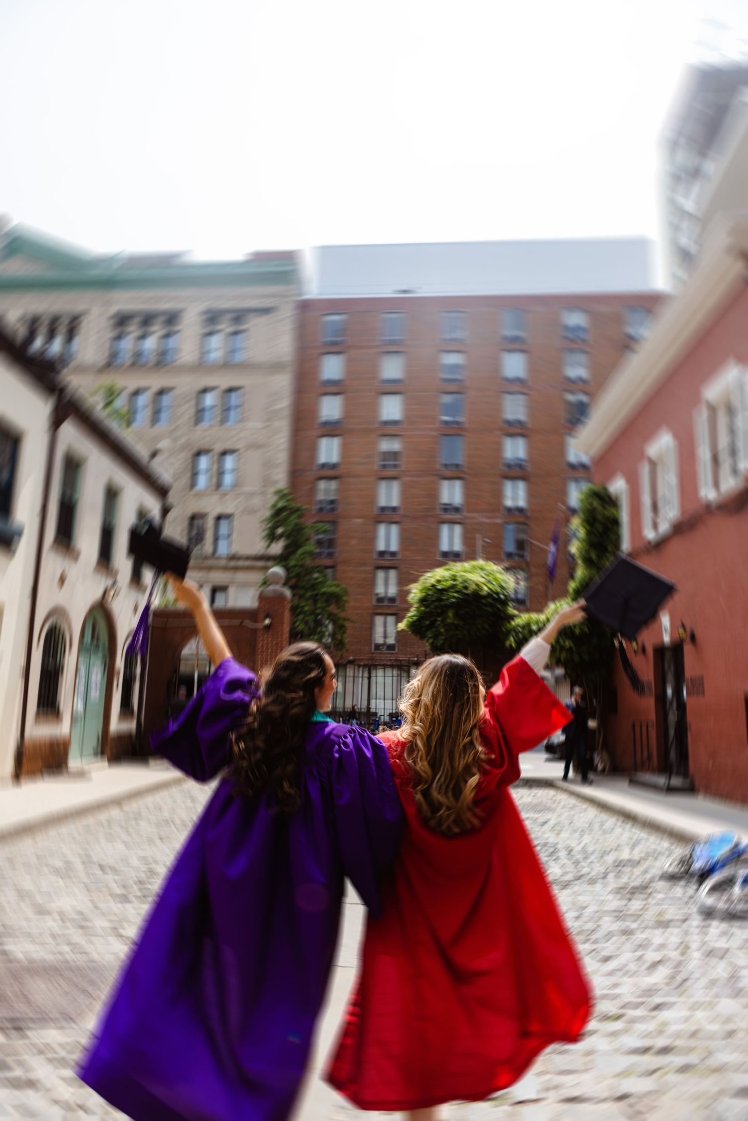 Two graduates in purple and red robes celebrate on a cobblestone street tossing caps in the air.