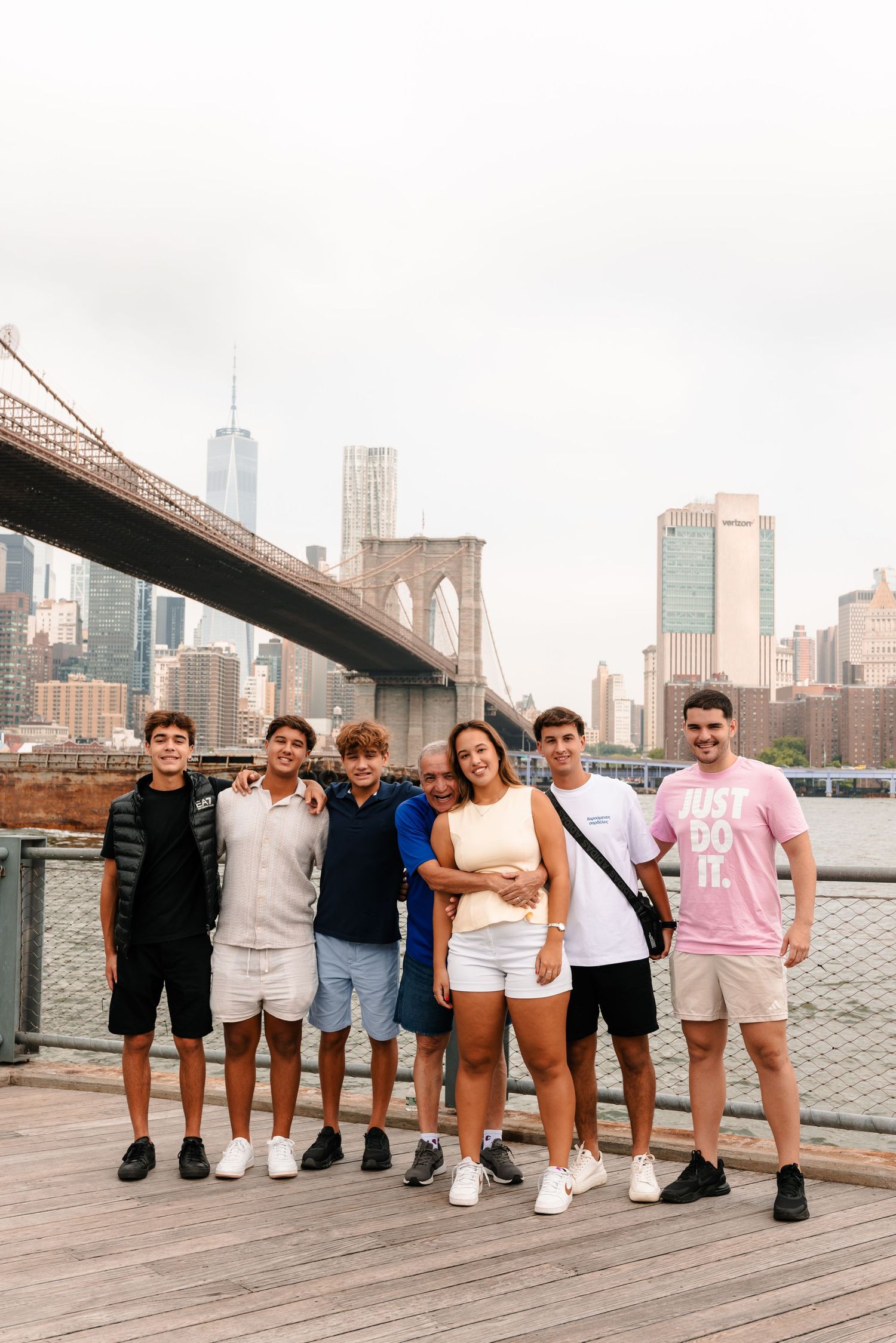 Group of seven young friends posing on wooden boardwalk with Brooklyn Bridge and Manhattan skyline behind them.