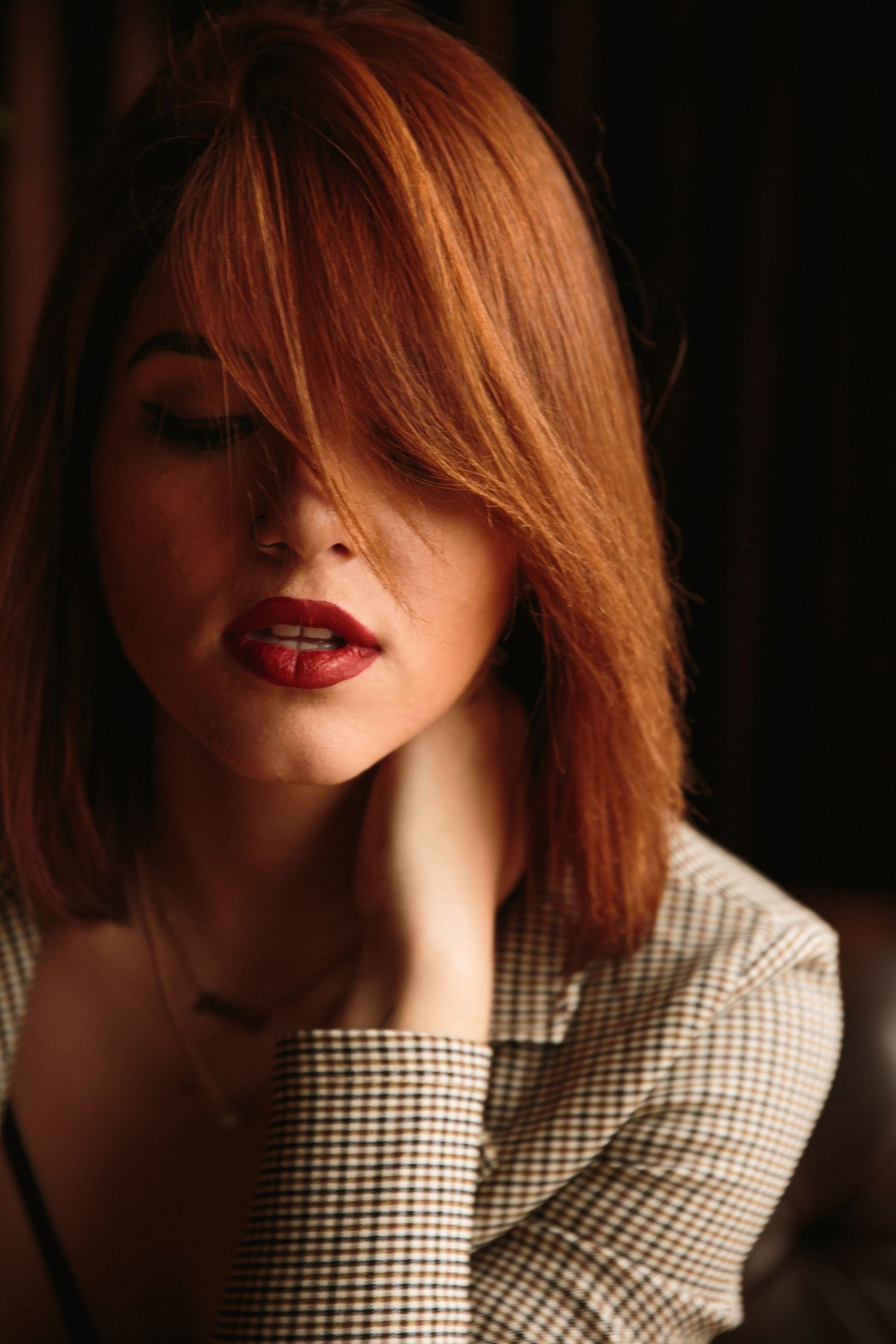 Moody portrait of a redhead woman in plaid blazer with hair covering one eye, red lips.