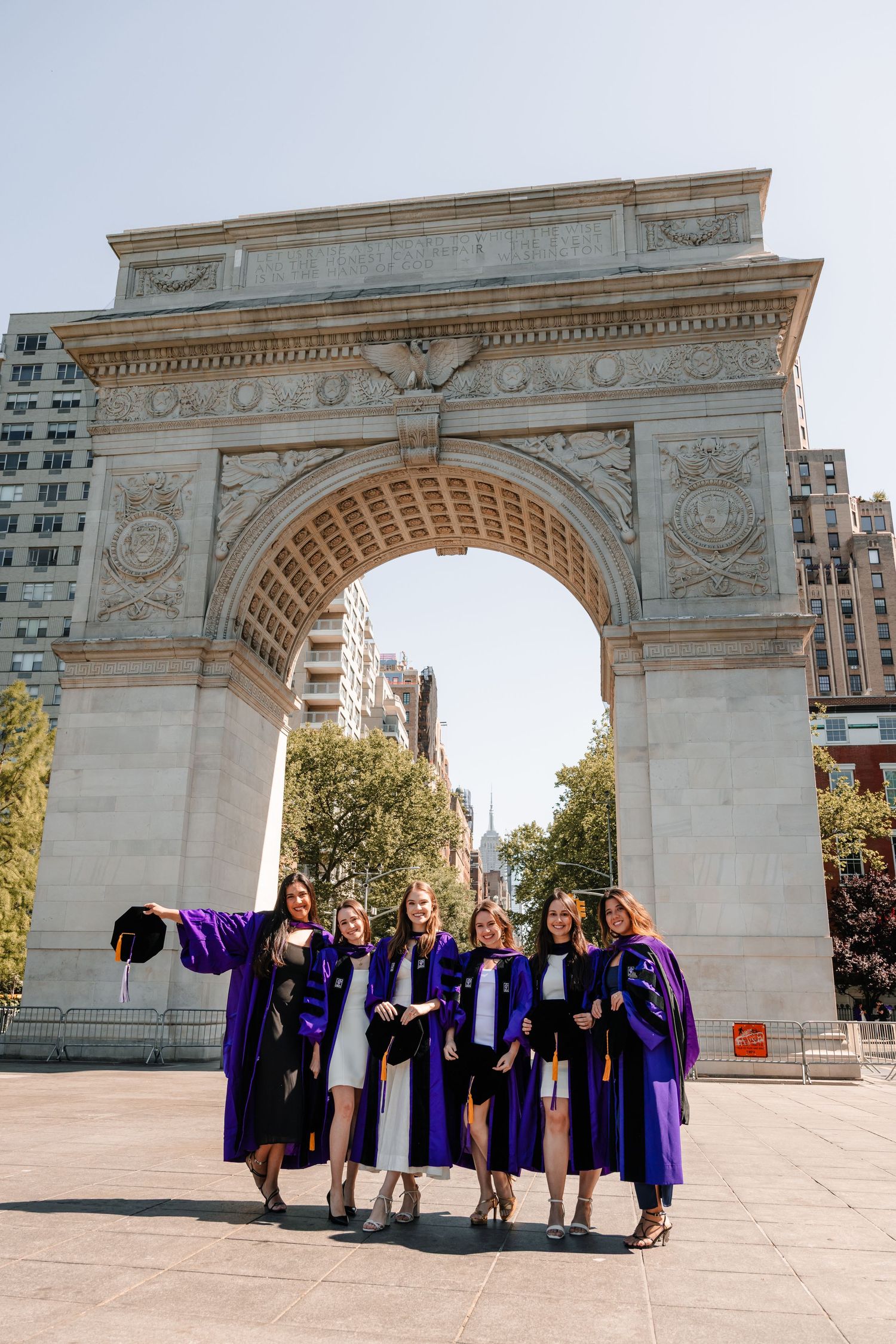 Group of NYU graduates in purple robes pose together at Washington Square Arch on sunny day.