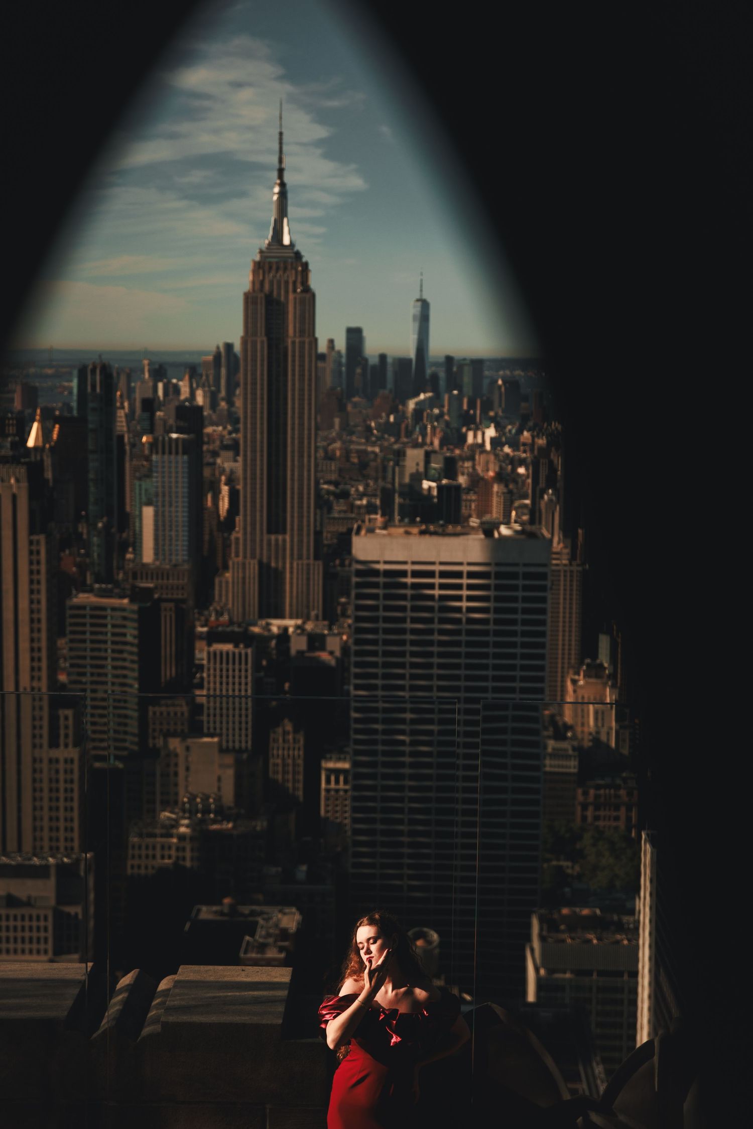 Woman in red dress standing on rooftop observation deck framed by arch with Empire State Building skyline view.