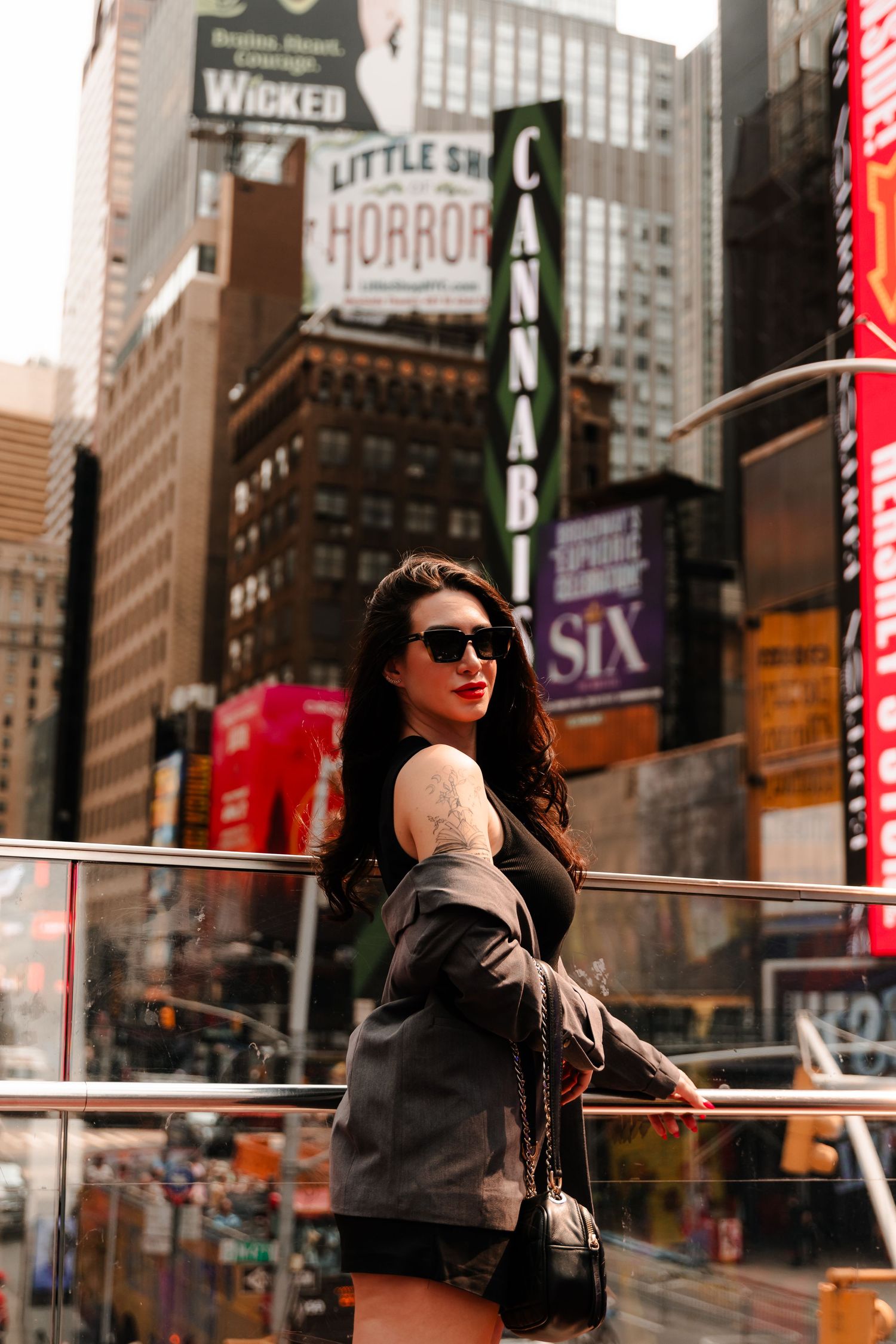 Brunette woman in sunglasses posing on Times Square balcony with Broadway signs behind her.
