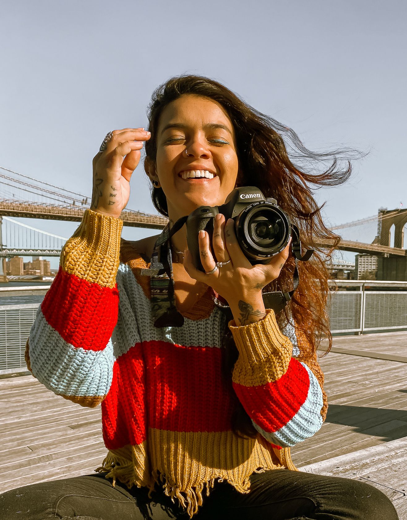 Joyful woman in multicolor knit sweater poses with Canon camera on sunny Brooklyn Bridge Park boardwalk.