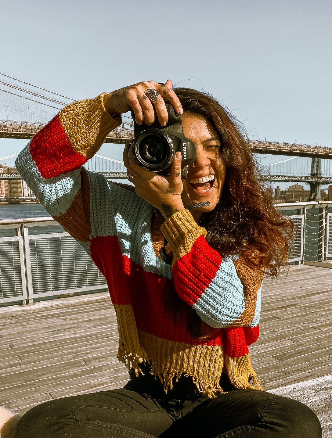 Laughing woman in colorful striped sweater holds Canon camera on Brooklyn waterfront with bridge backdrop.