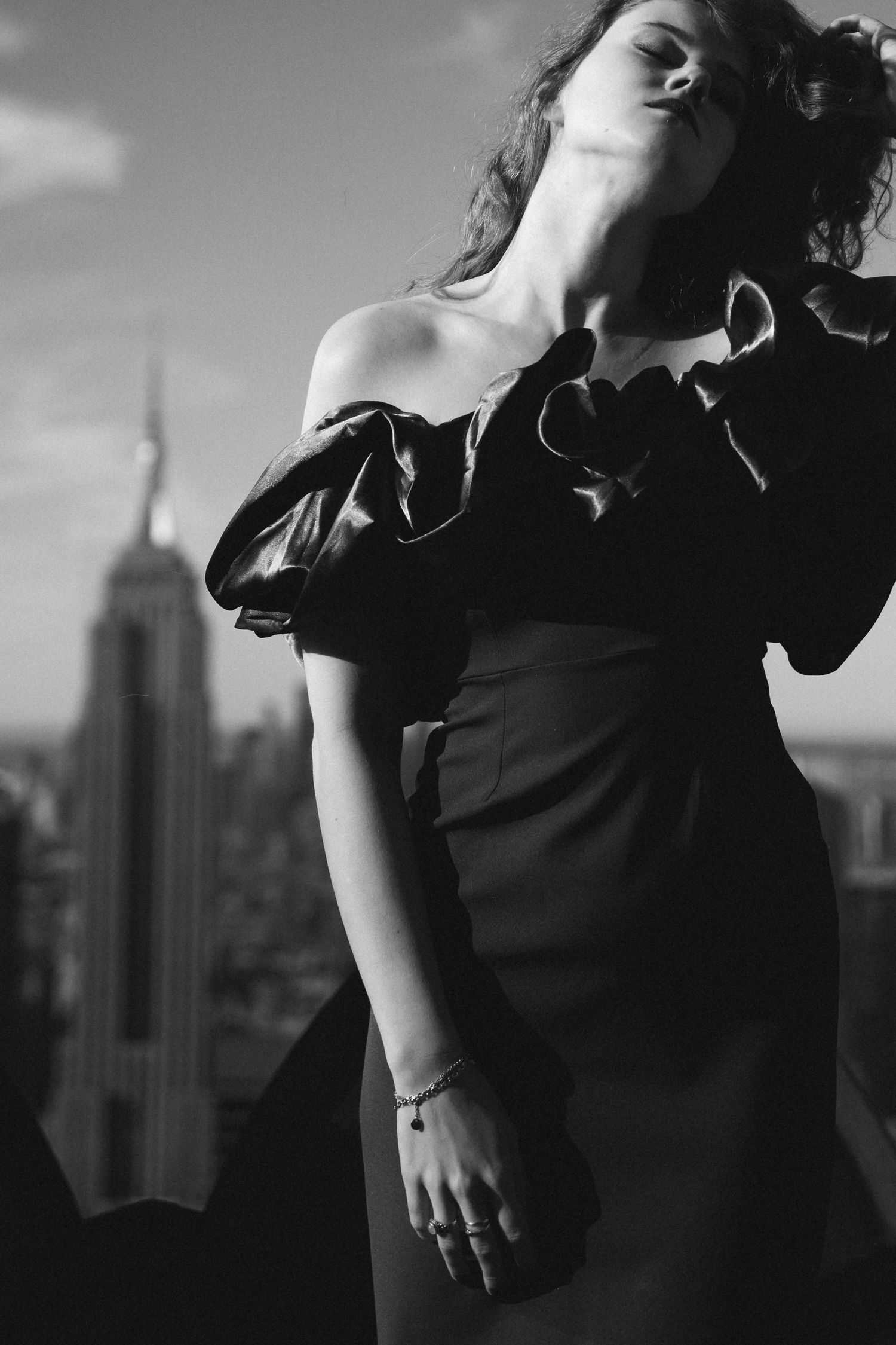 Black and white portrait of woman in off-shoulder ruffled dress with Empire State Building skyline in background.