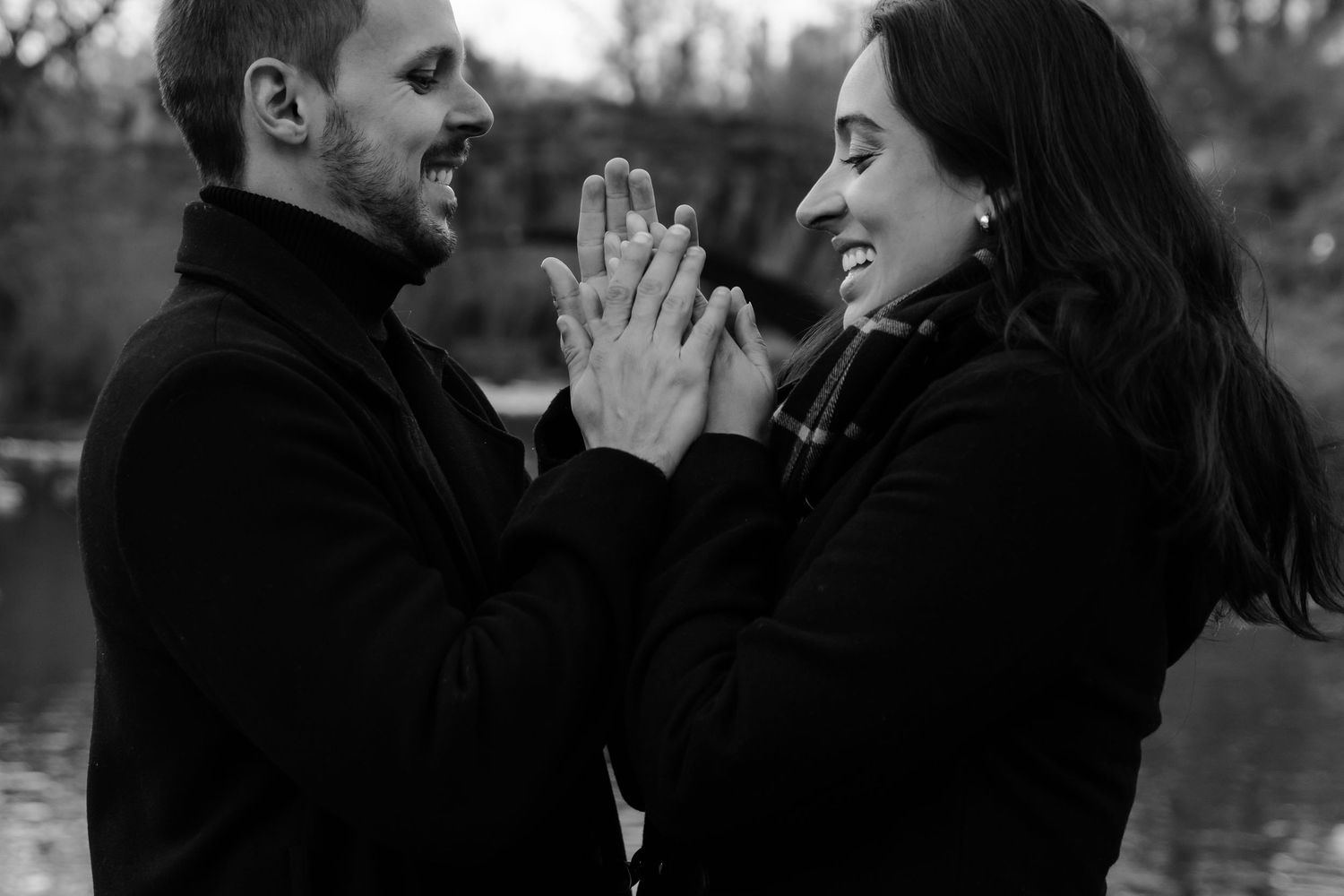 Black and white photo of smiling couple pressing palms together near a lake in Central Park.