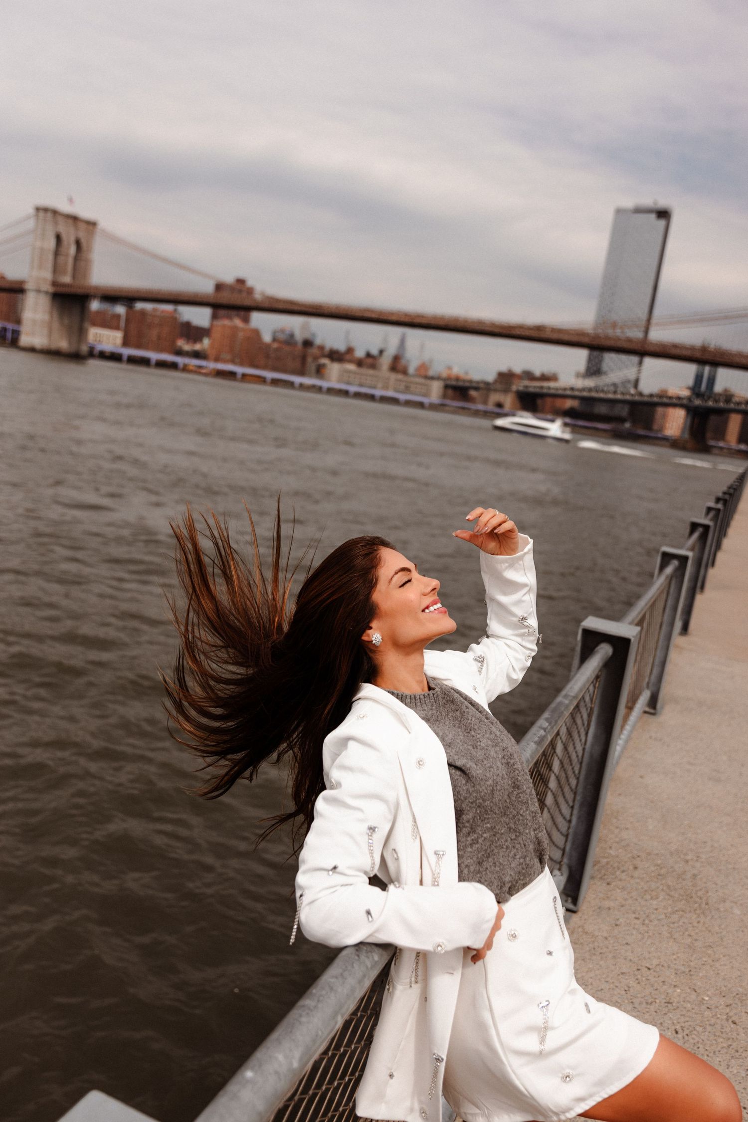 Joyful woman tossing hair by Brooklyn Bridge waterfront wearing white jacket and gray dress.