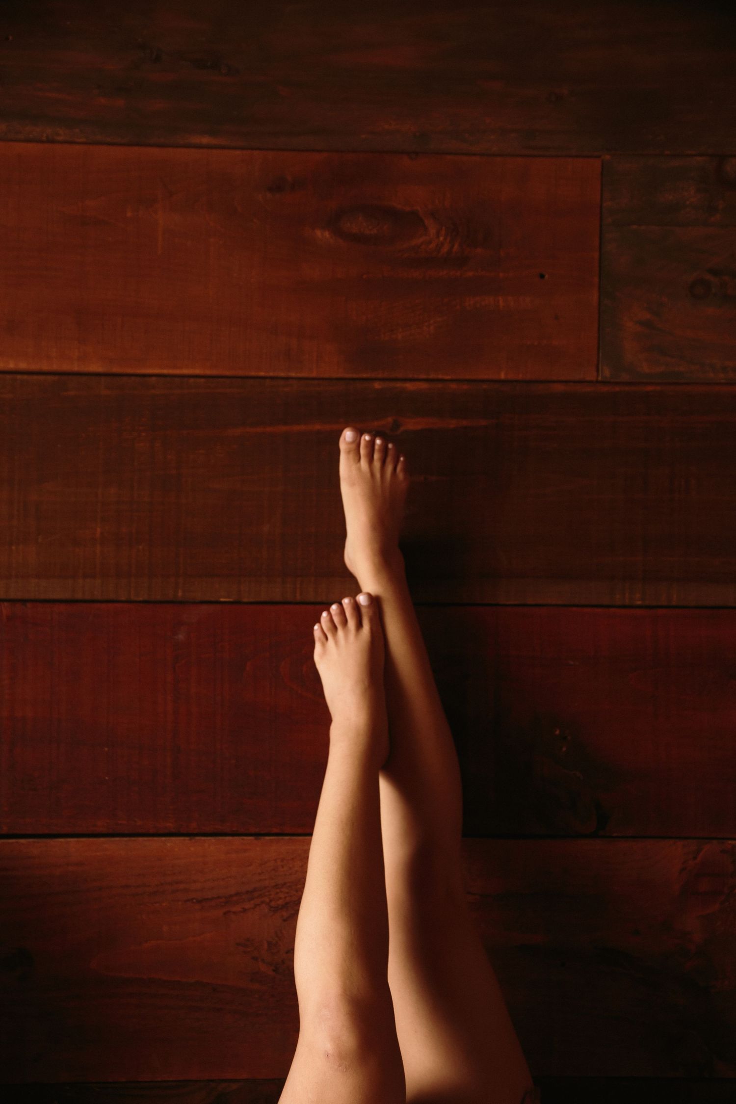 Bare feminine legs crossed at ankles resting on rich dark hardwood floor in soft lighting.