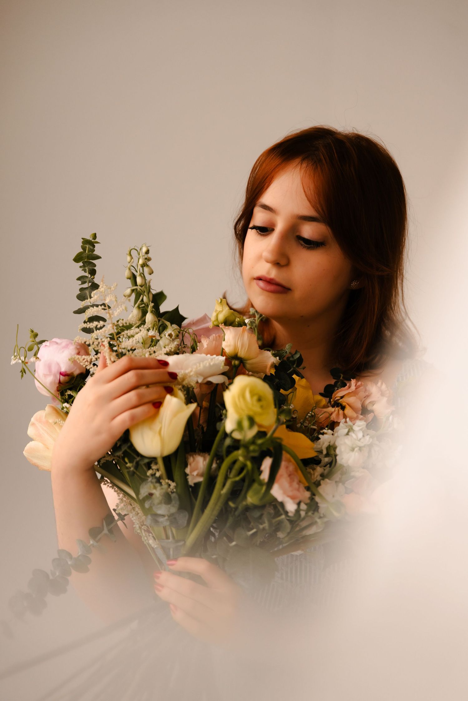 Young redhead woman in white dress gently holds a lush bouquet of mixed flowers with eyes cast downward.