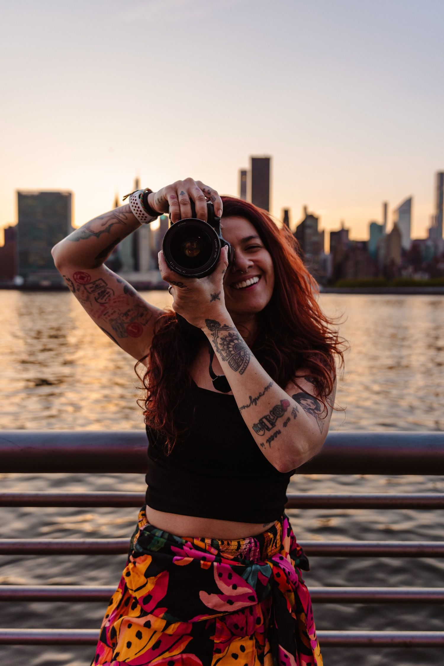 Smiling tattooed woman photographing with camera against NYC skyline at sunset.