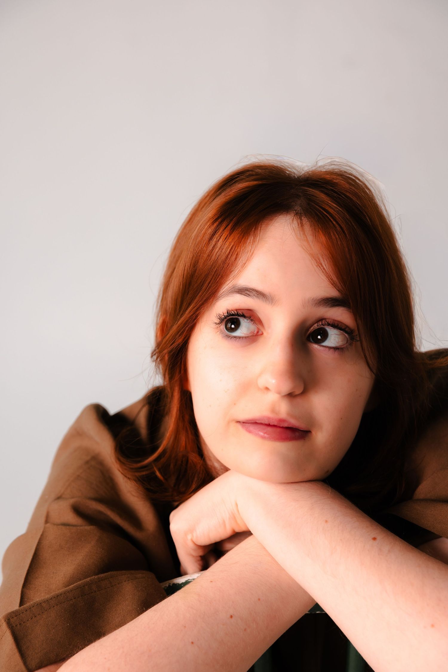 Pensive young redhead woman rests chin on folded hands while looking away dreamily against a light gray backdrop.