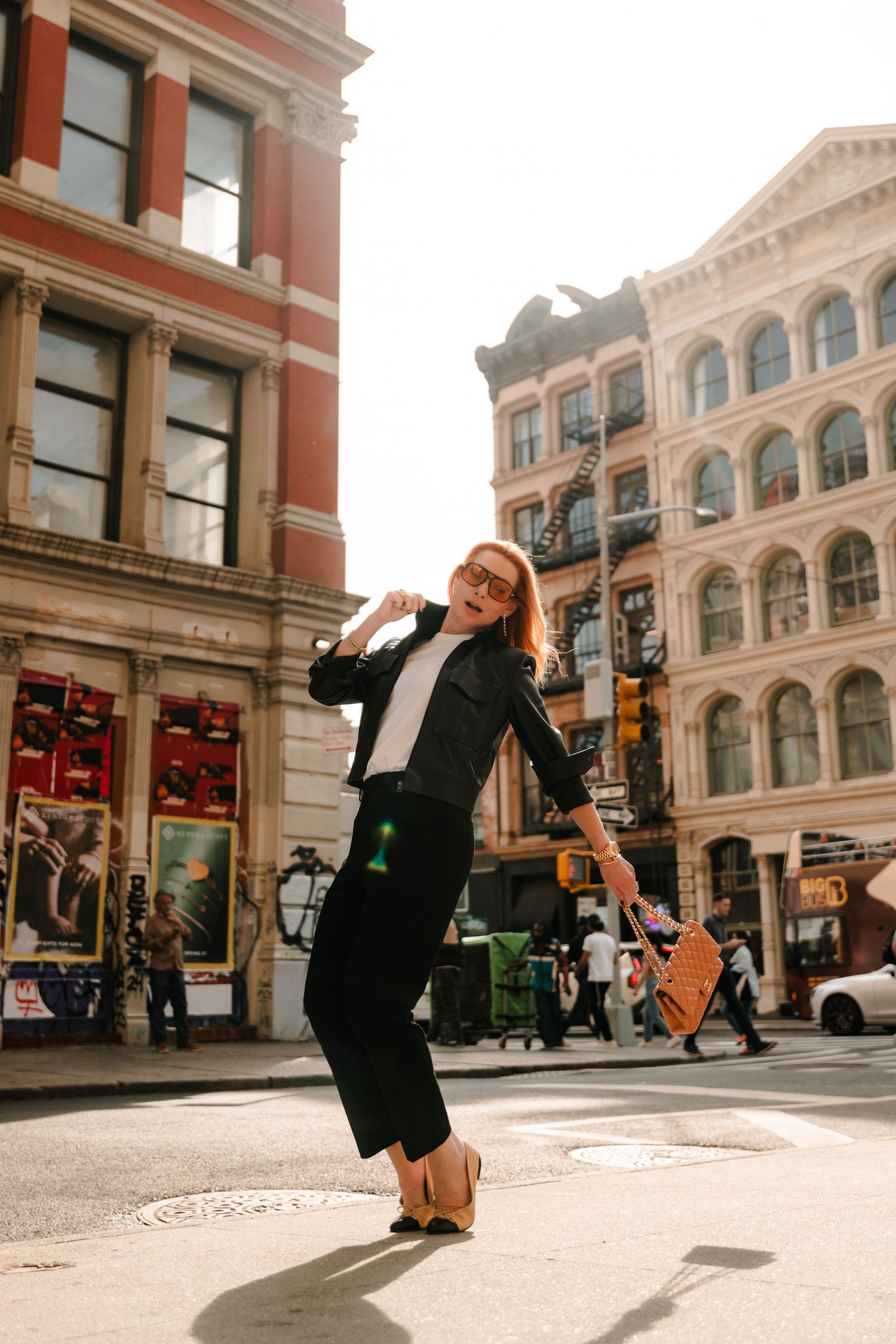 Redhead woman in black leather jacket and pants poses playfully on sunny New York City street corner.