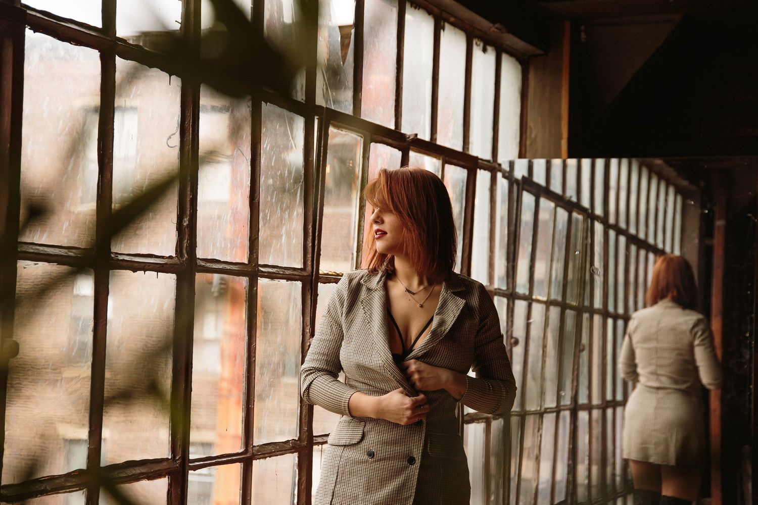 Redhead woman in grey plaid suit standing by vintage industrial windows with reflection.