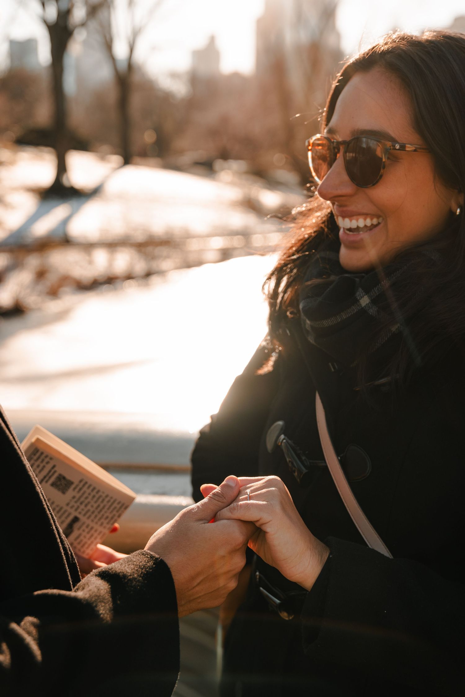 Smiling woman in sunglasses holds hands with partner on a sunny winter day in Central Park.