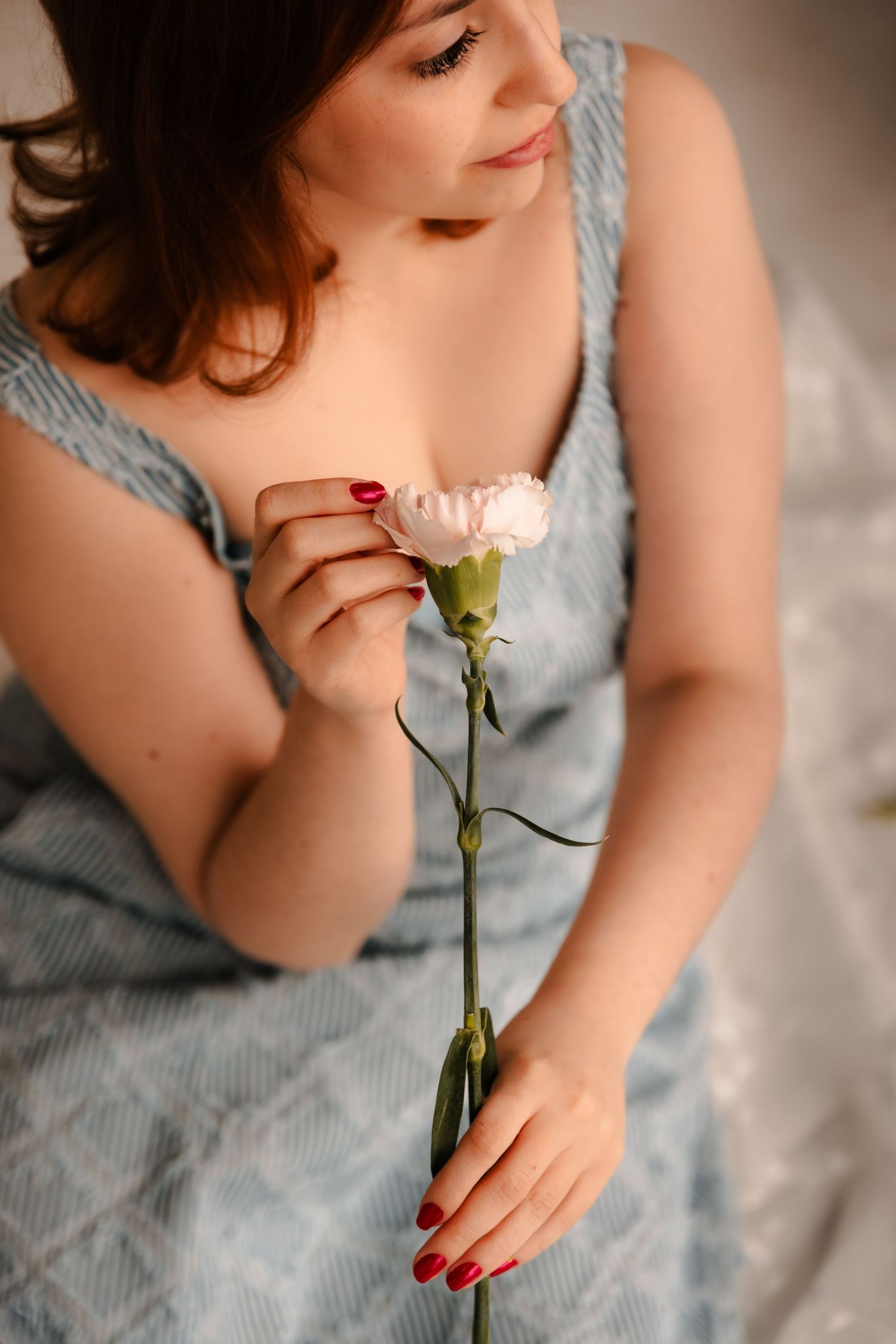 Young woman in blue striped dress sits on bed holding a single delicate white carnation with red-painted nails.