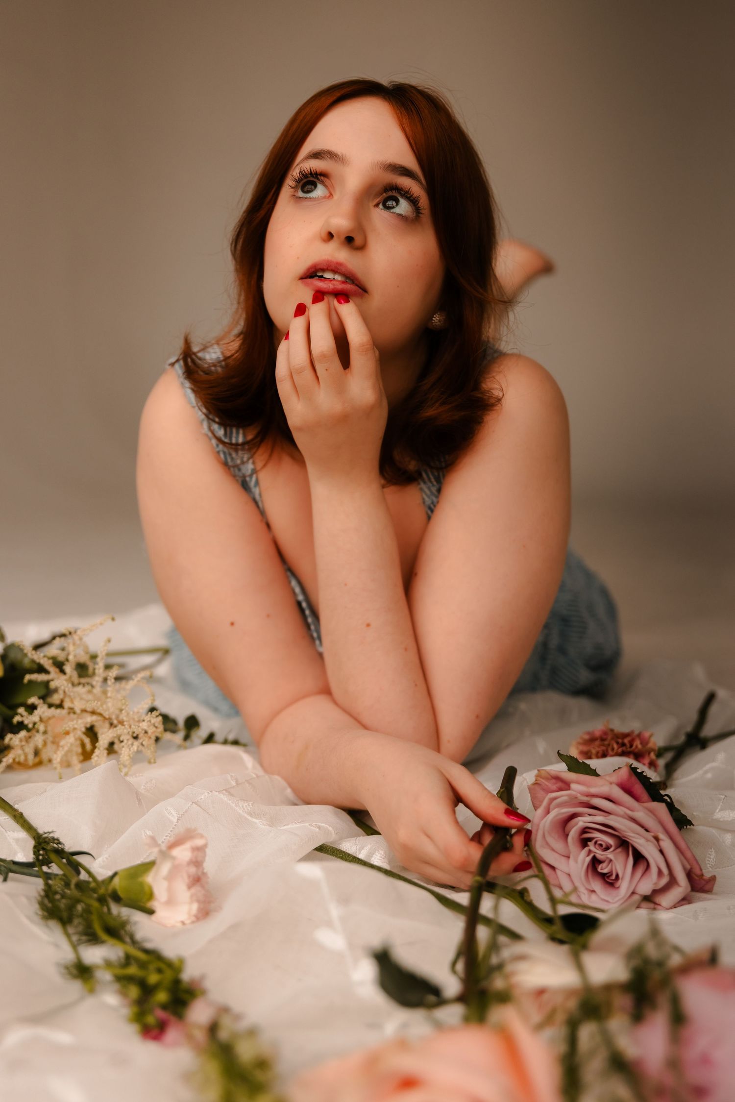 Young woman with red nails lies among scattered roses and flowers on white fabric, gazing upward dreamily.
