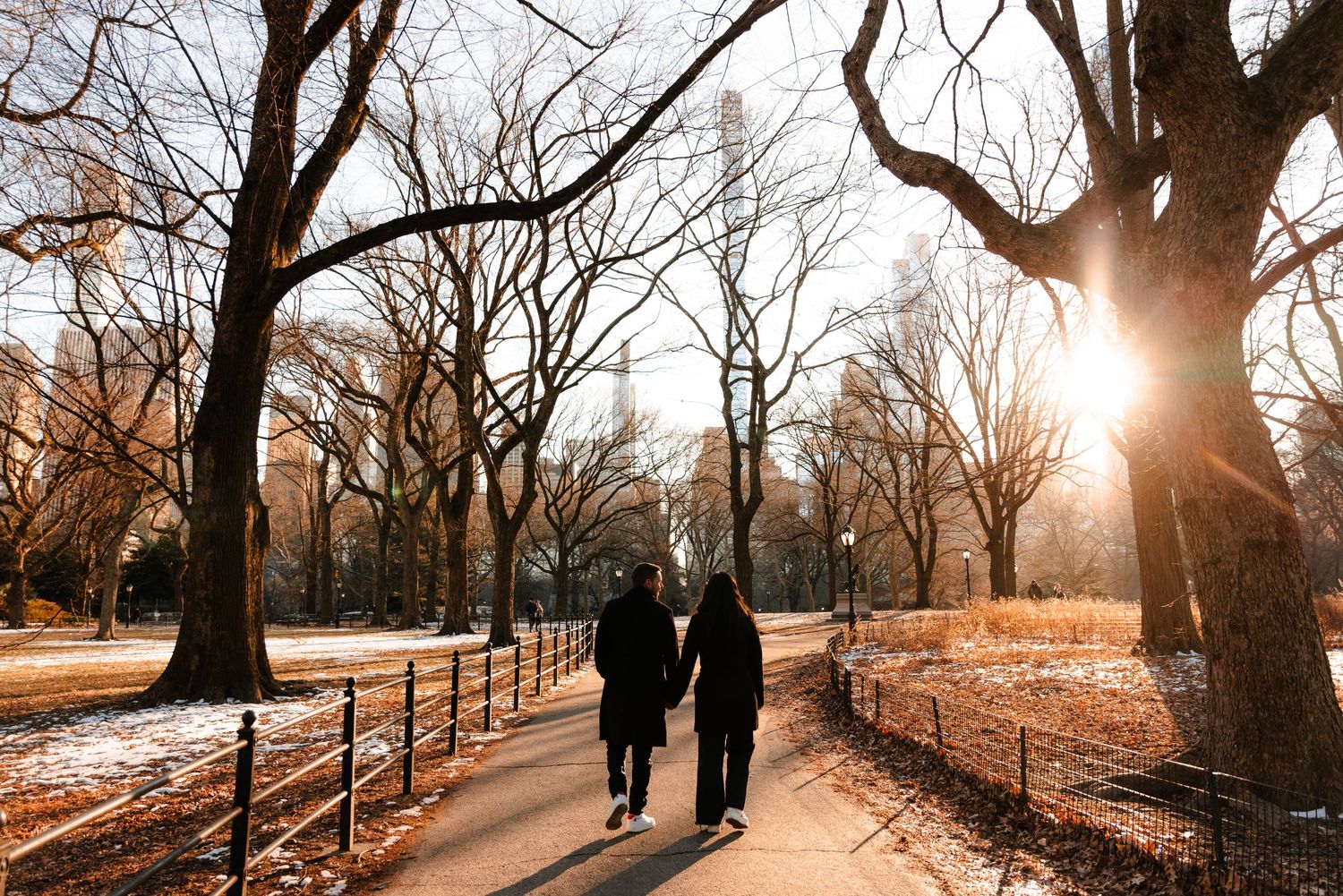 Couple walks hand in hand along a sunlit Central Park path surrounded by bare winter trees.