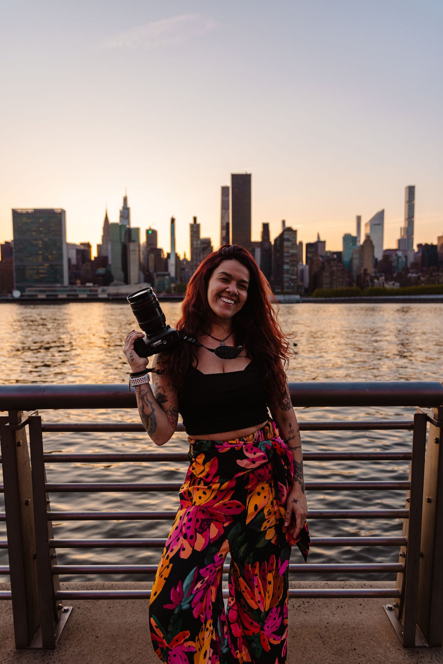 Tattooed woman with red hair holding camera at NYC waterfront sunset.