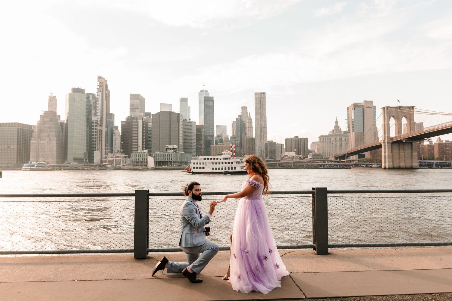 Man proposes on one knee to woman in lavender gown with NYC skyline and Brooklyn Bridge backdrop.