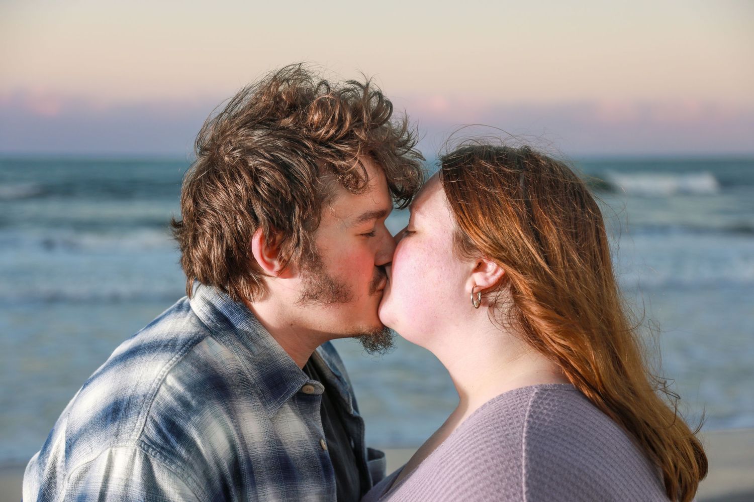 A young couple shares a romantic kiss on the beach at sunset with ocean waves in the background.