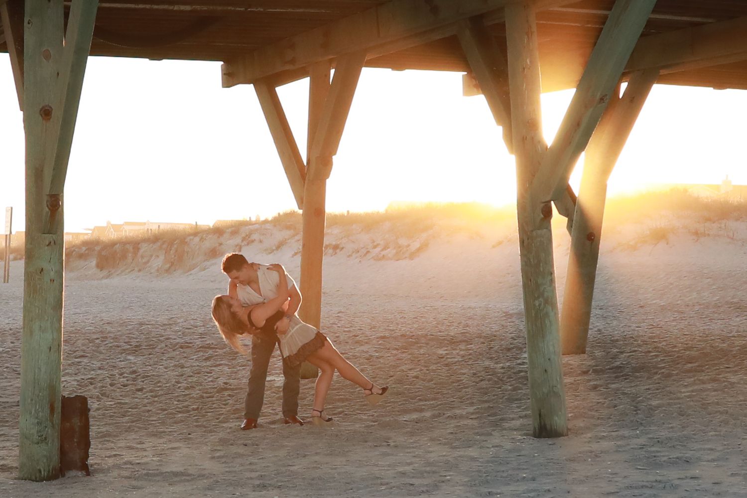 Couple embracing romantically under a wooden pier on a sandy beach at golden sunset.