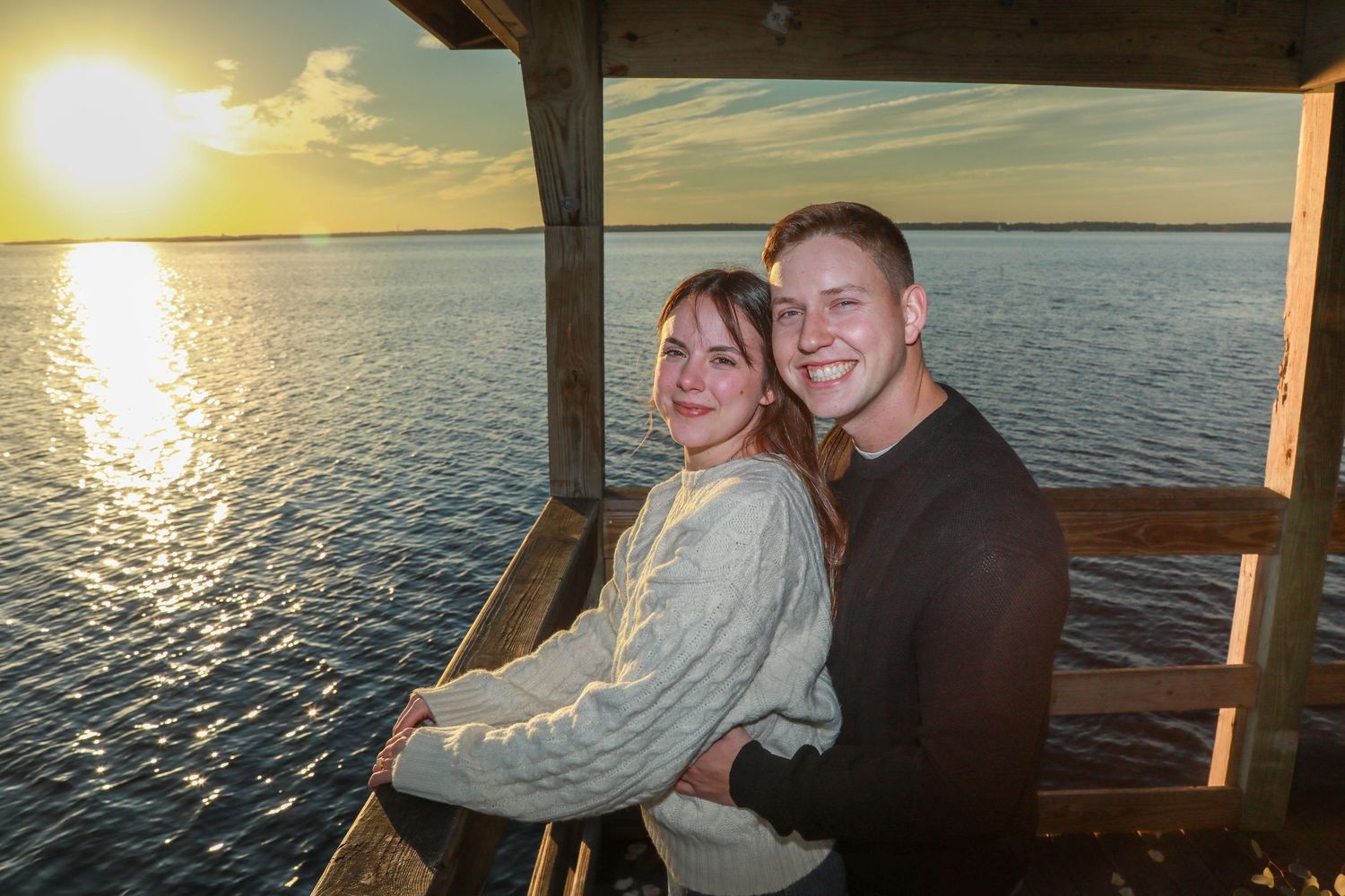 Happy couple embracing on a wooden dock at golden hour sunset over calm water.
