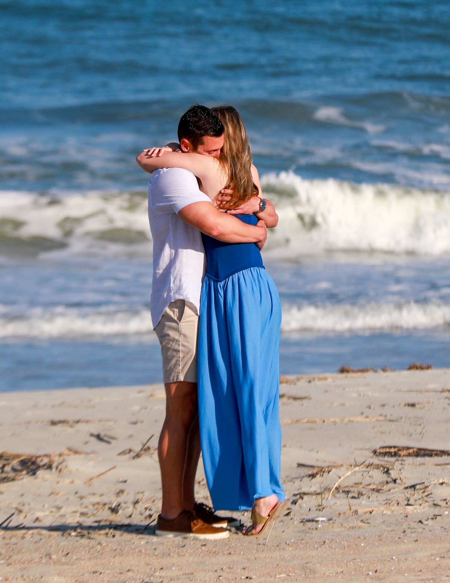Couple embracing romantically on a sandy beach, man in white shirt and woman in blue maxi dress, ocean waves in background.