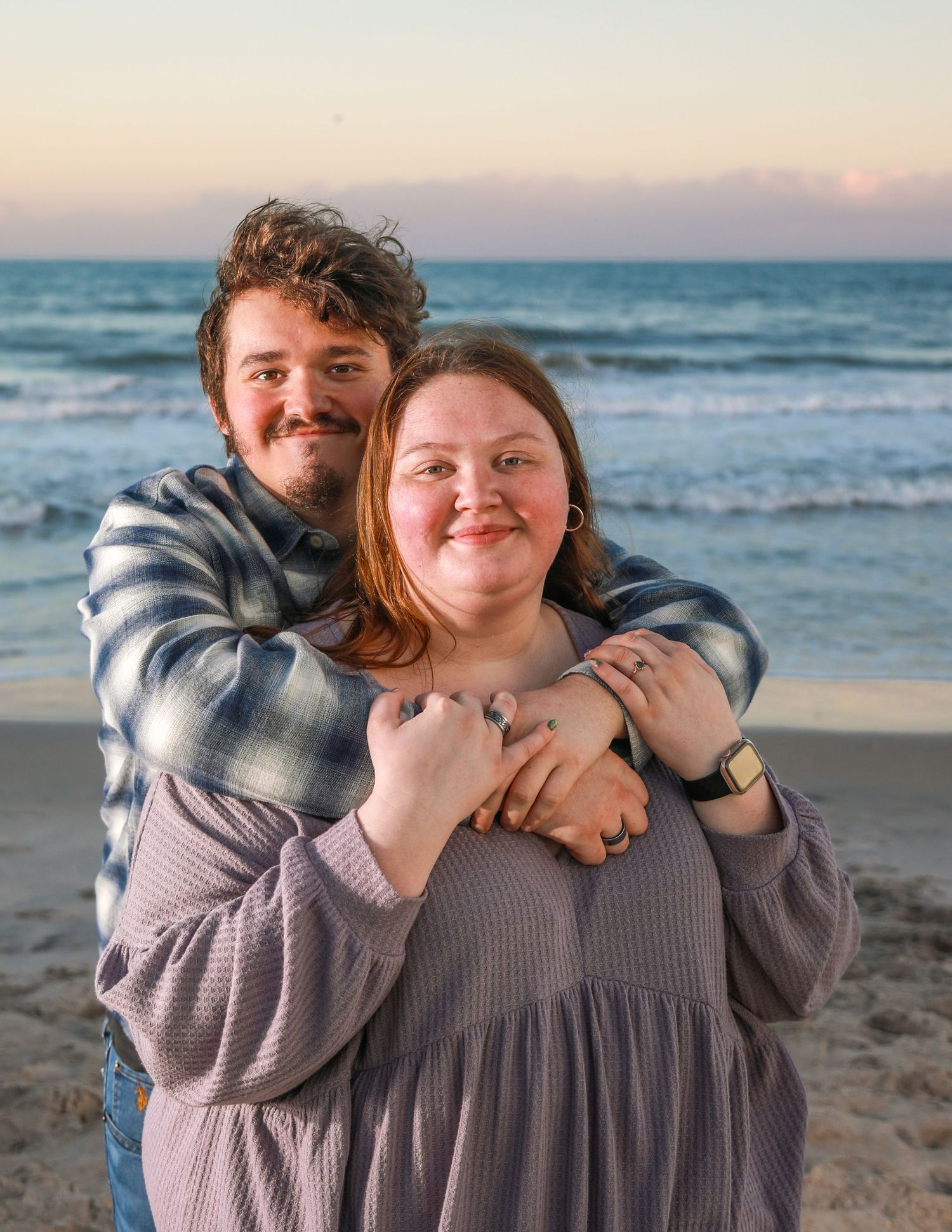 Couple embracing on a beach at sunset, smiling warmly with ocean waves in the background.