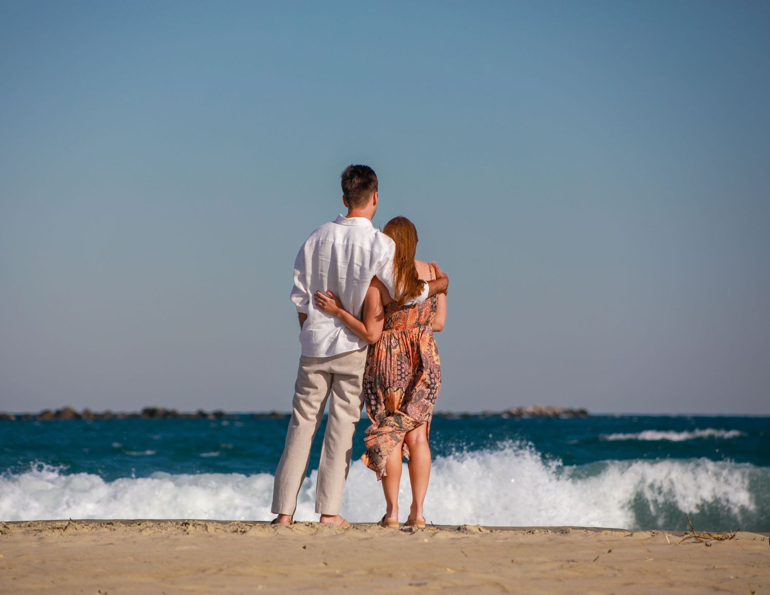 Couple standing together on a sandy beach, embracing while watching ocean waves under a clear blue sky.