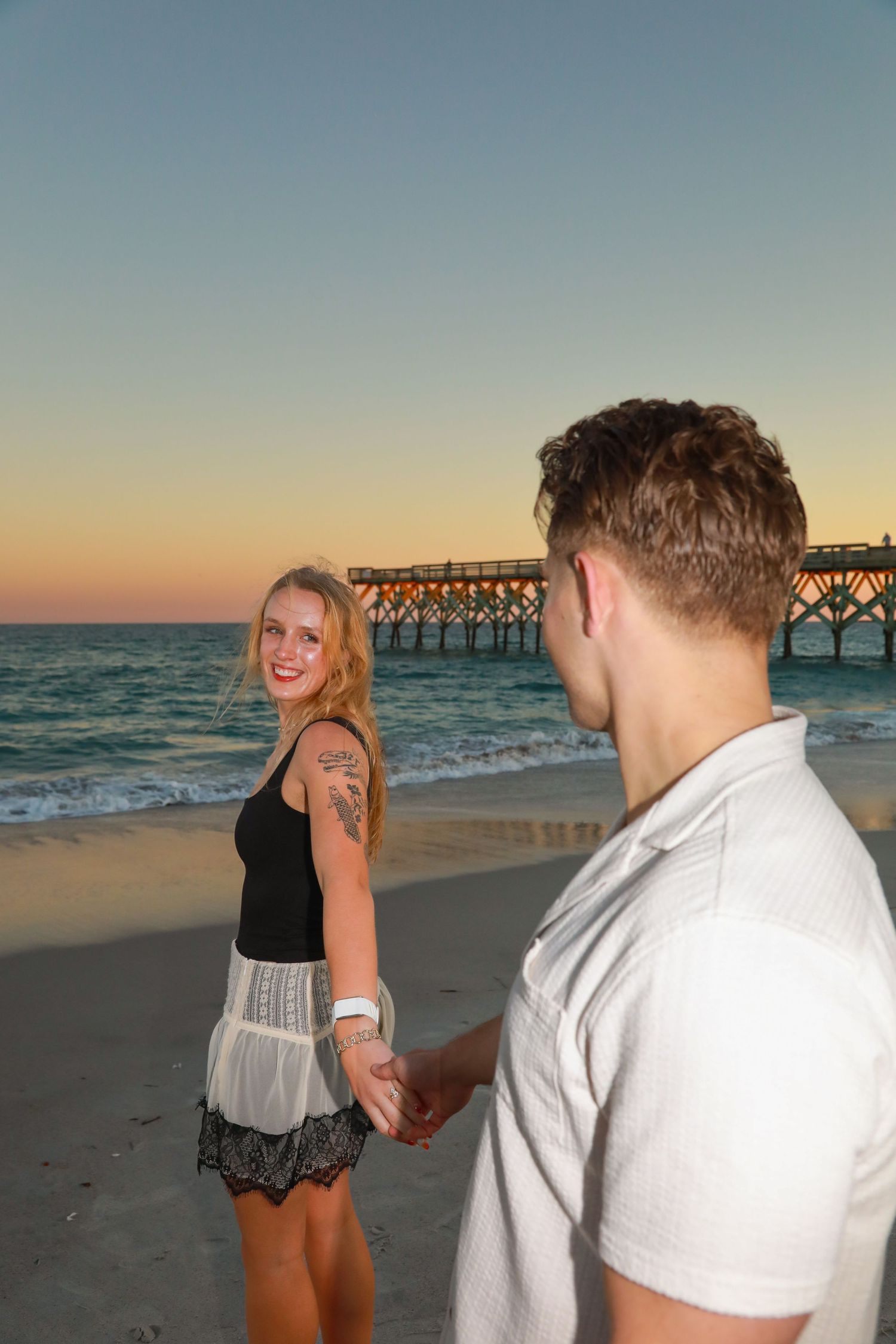 Couple holding hands on a beach at sunset with a pier in the background, woman smiling in black top and lace skirt.