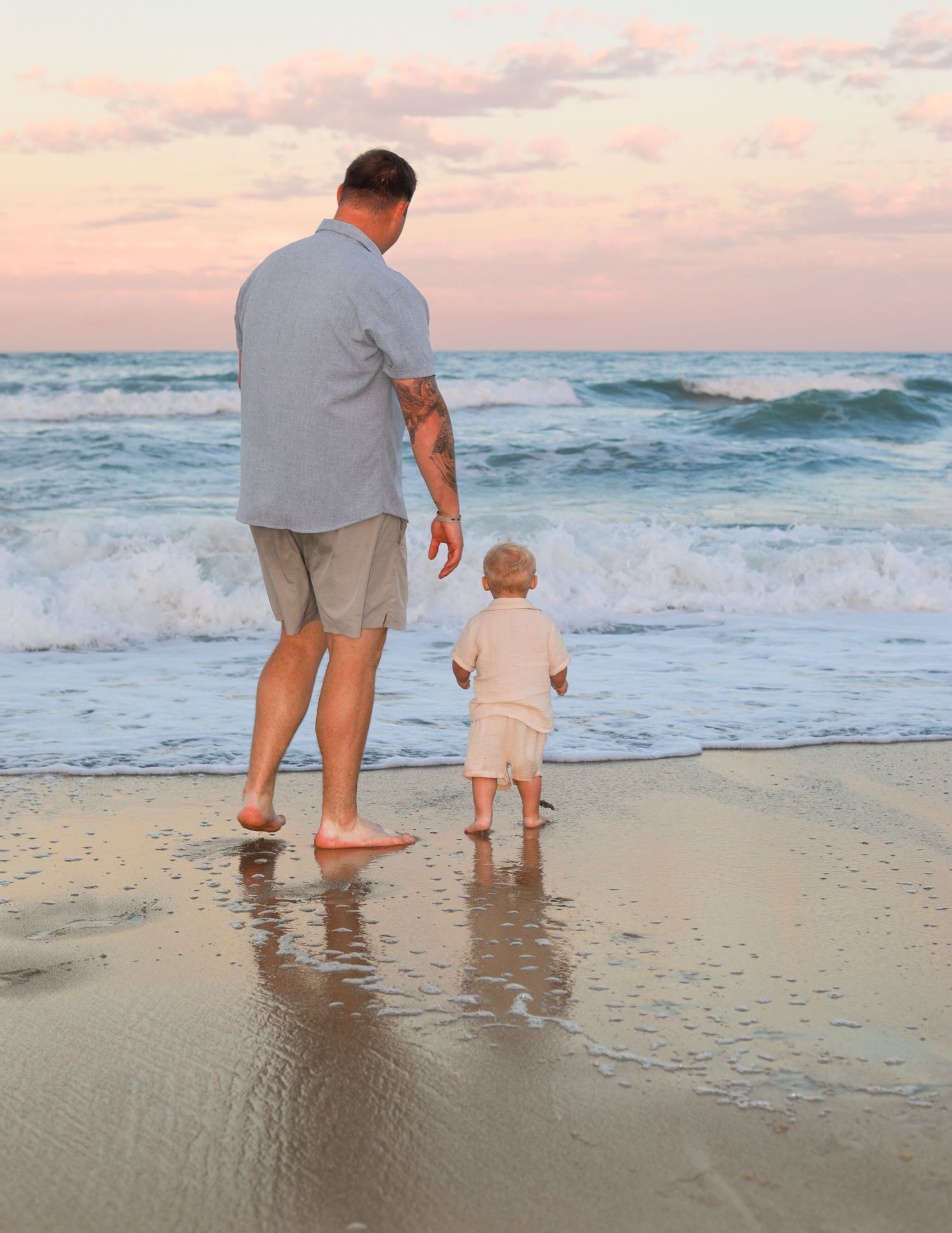 Father and toddler standing barefoot on a sandy beach at sunset, watching gentle ocean waves together.