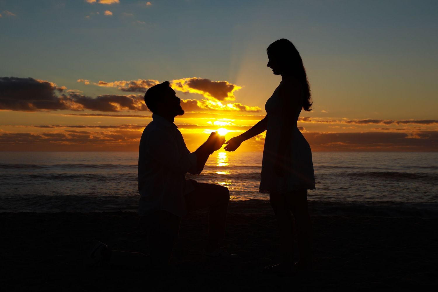 Silhouette of a man proposing on one knee to a woman on a beach at sunset with glowing orange sky.