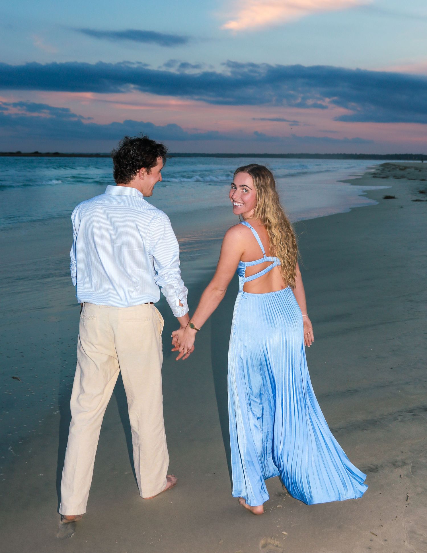 Couple holding hands on a beach at sunset, woman in blue pleated dress, man in white shirt and khaki pants.