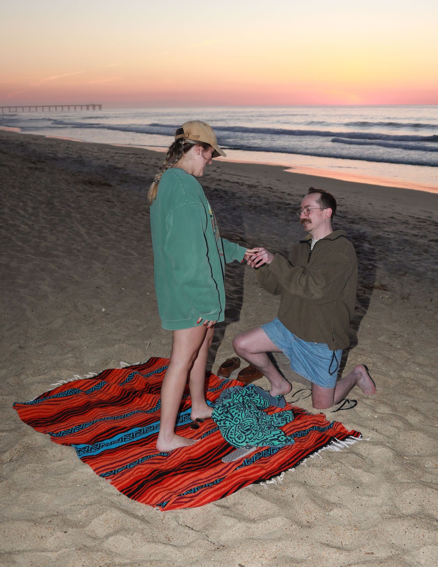 Man proposes to woman on a colorful blanket at a beach during a beautiful sunset with a pier in the background.