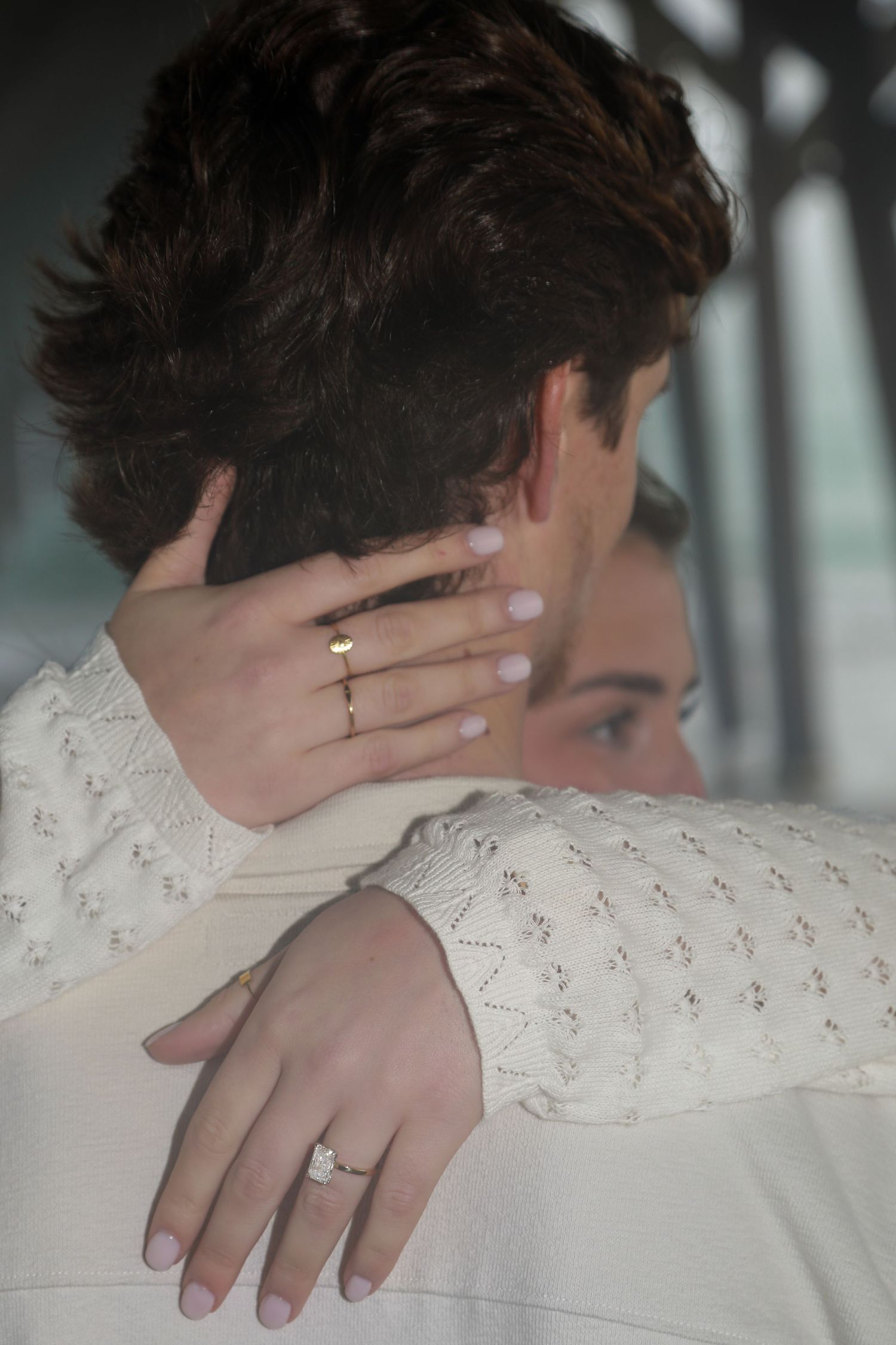 Couple embracing, woman wearing diamond engagement ring and gold band on her hand, dressed in white knit sweater.